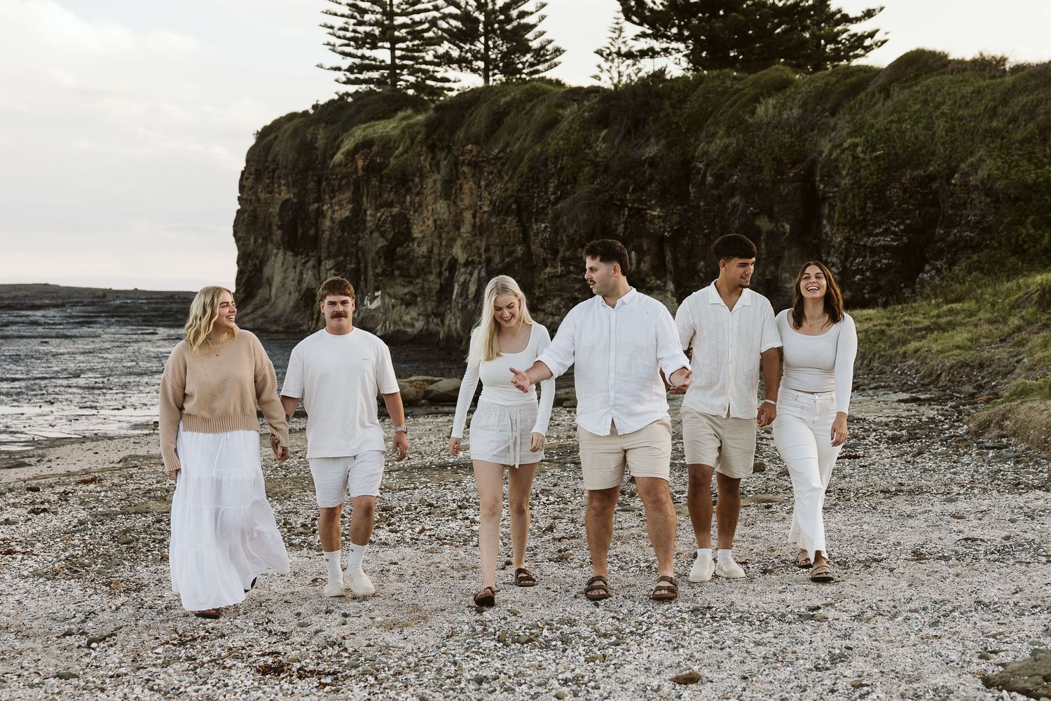 Family of six walking on a rocky beach near coastal cliffs, dressed in coordinated white and beige outfits.
