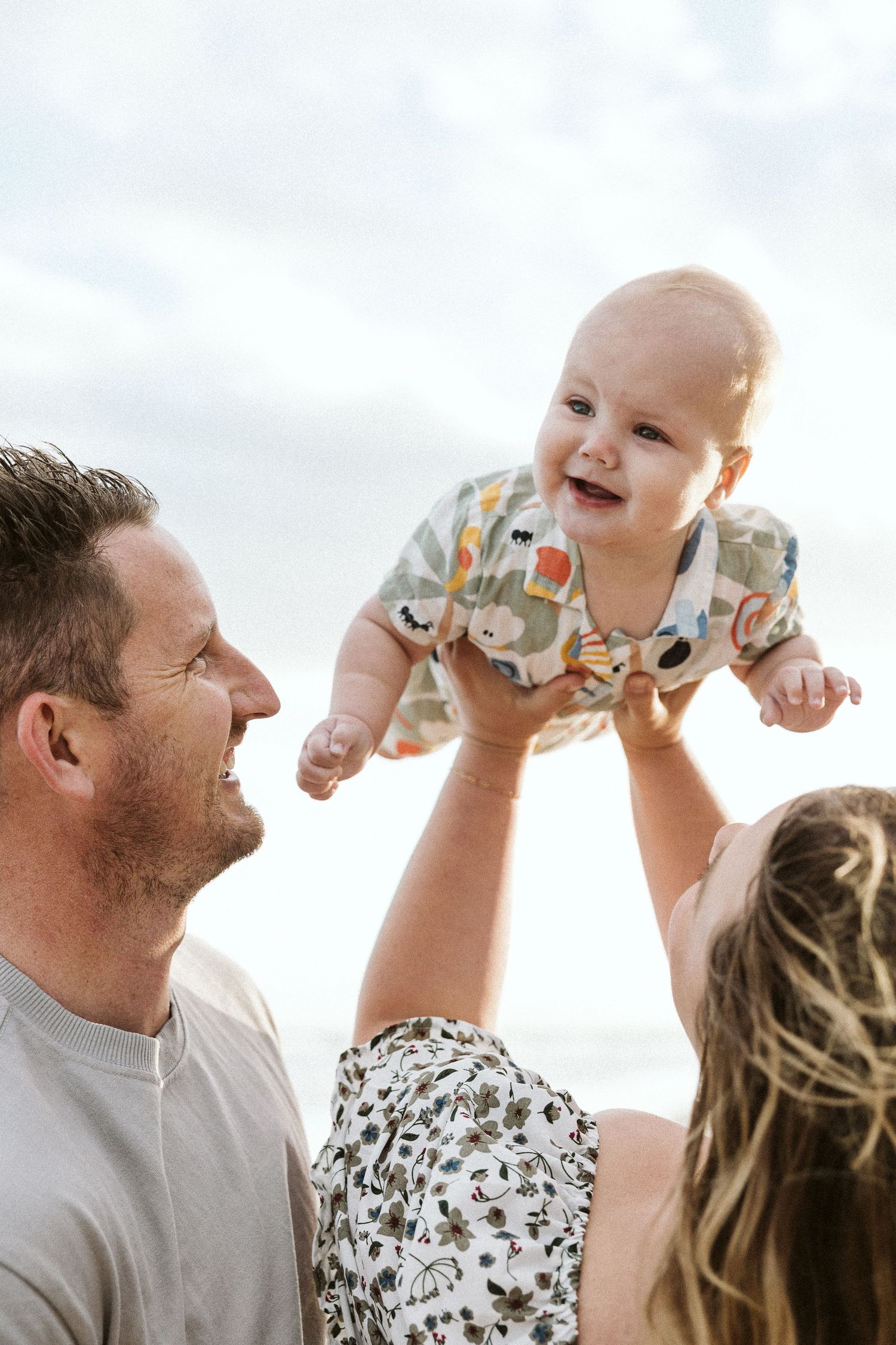 Happy baby lifted in the air by smiling parents outdoors under a cloudy sky.