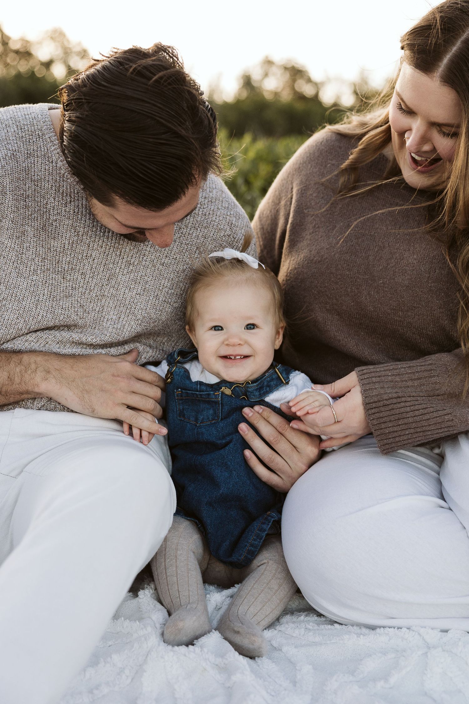 A happy baby in denim overalls sits between smiling parents on a white blanket outdoors in warm golden light.