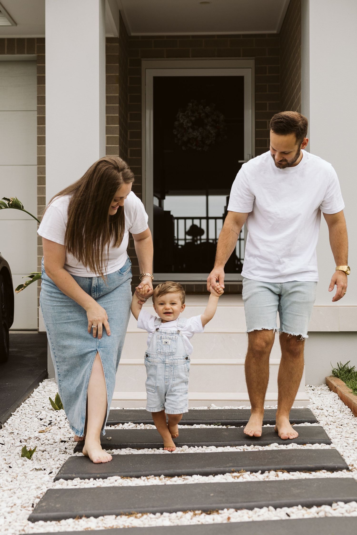 A young family holds hands with their smiling toddler on front steps of a modern home.