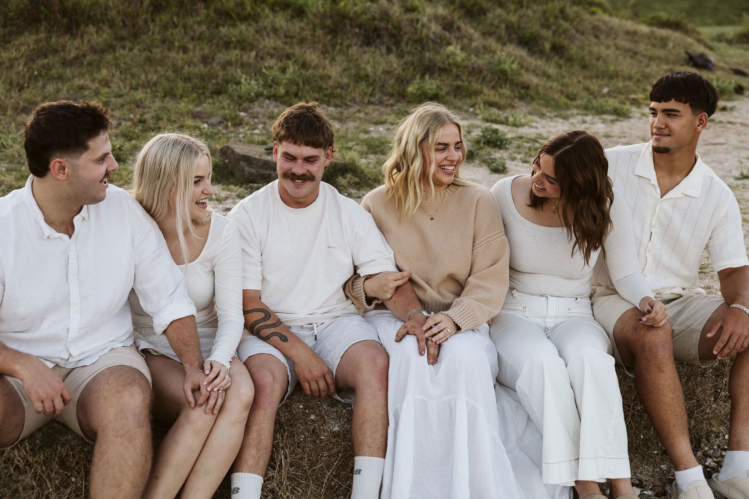 Six young friends laughing and sitting together outdoors on a sunny day, dressed in casual white and neutral tones.