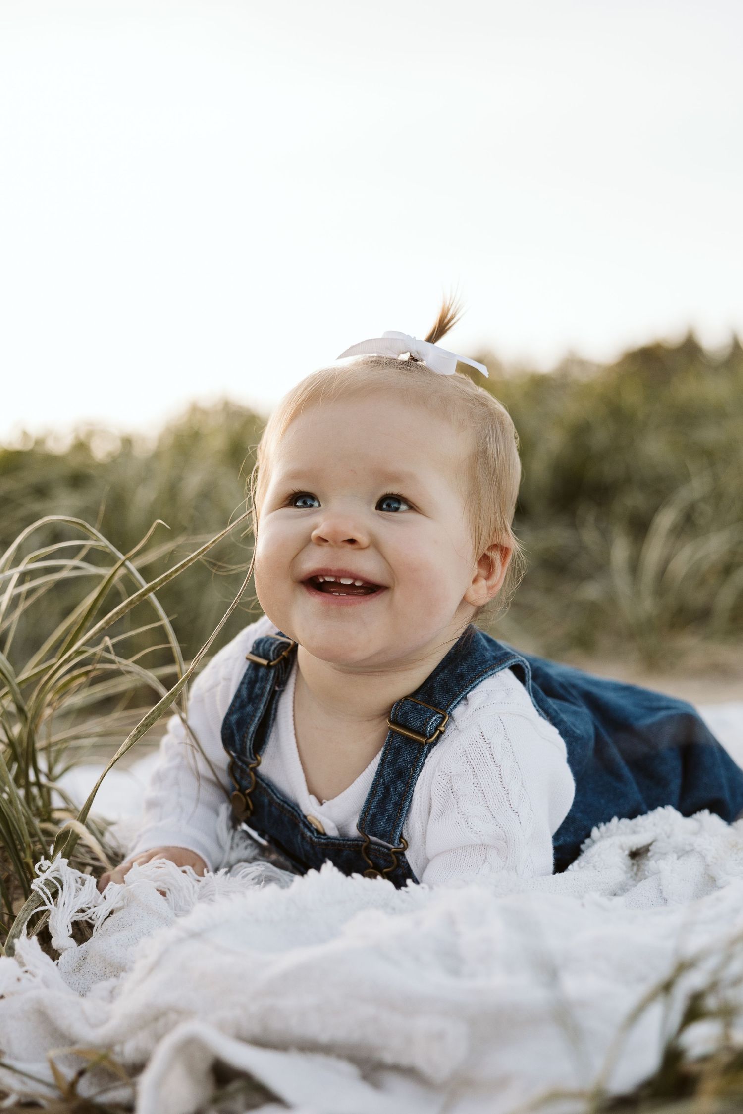 Smiling baby girl in denim overalls and white bow sits on blanket outdoors near green grass in golden light.
