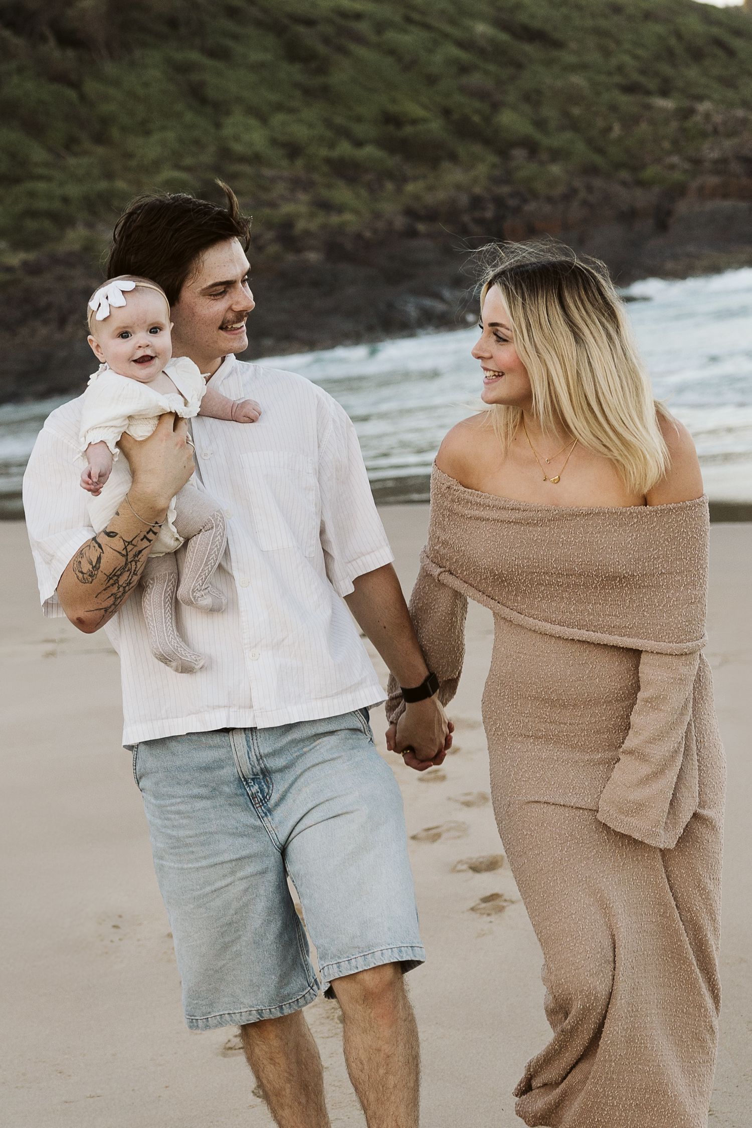 A young couple walks hand-in-hand on a beach, the man holding a baby girl, both smiling at each other.