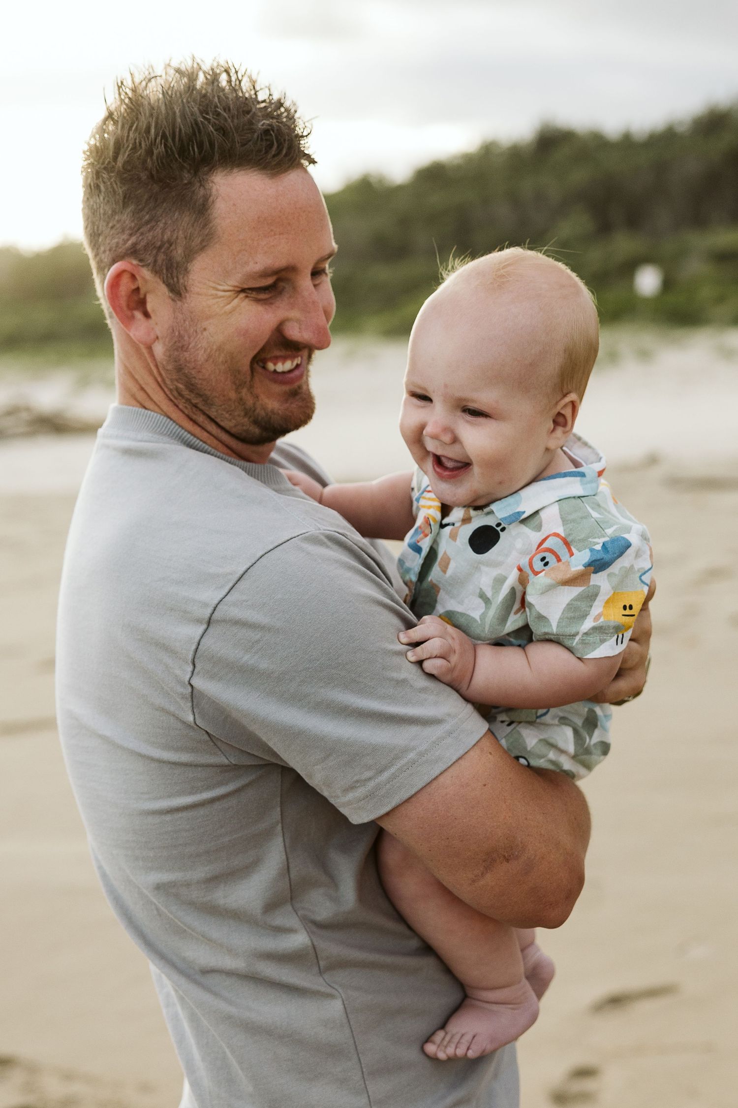 A smiling man holds a laughing baby on a sandy beach, sharing a joyful moment together outdoors.