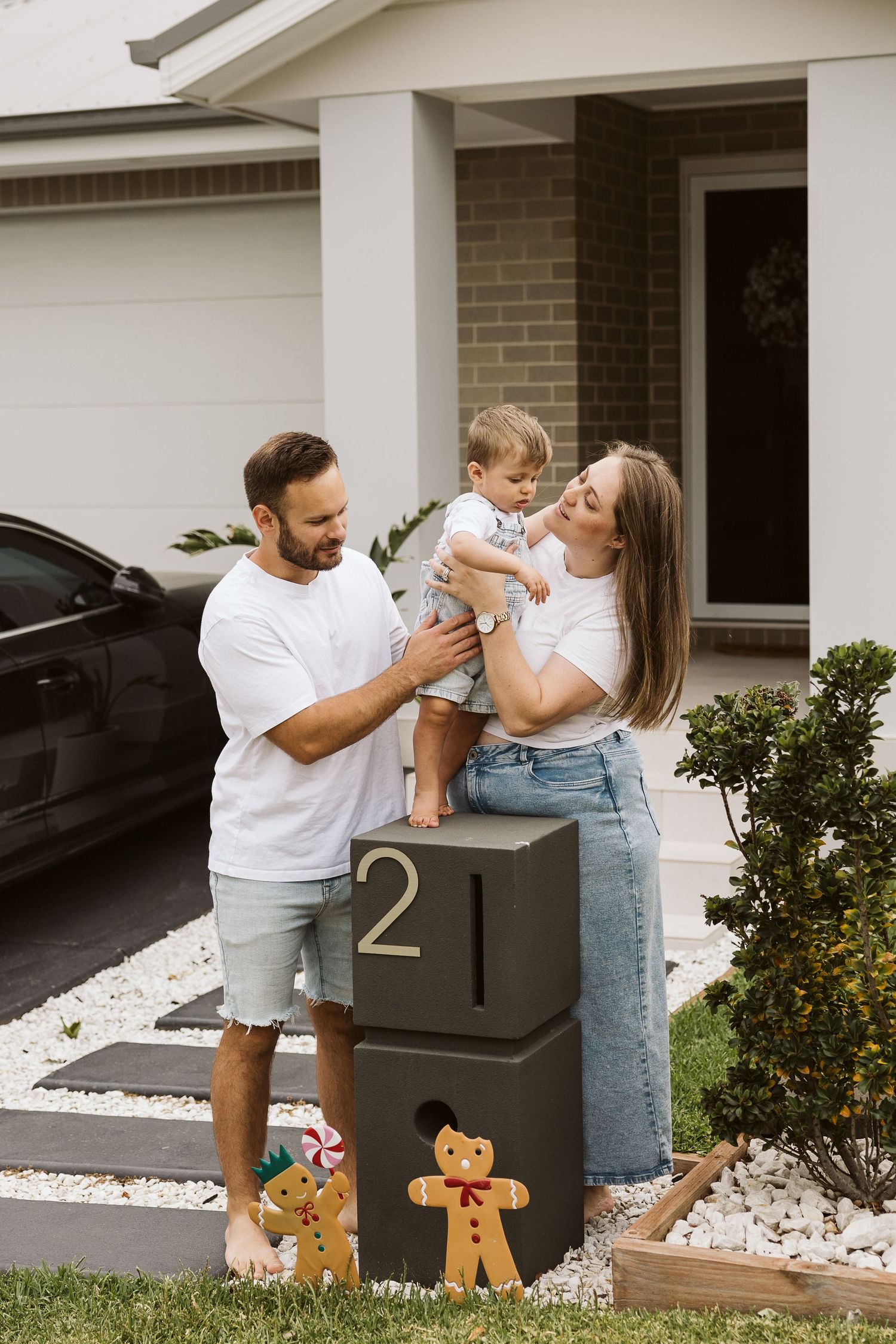 Happy couple holds their baby by a house number post with Christmas decorations outside.