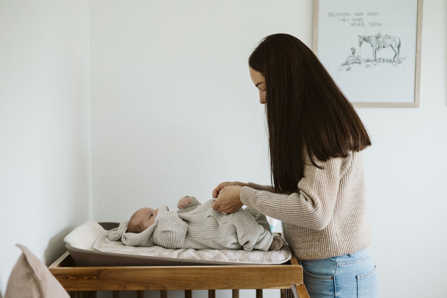 Young mother in beige sweater tends to her newborn baby on a changing table.