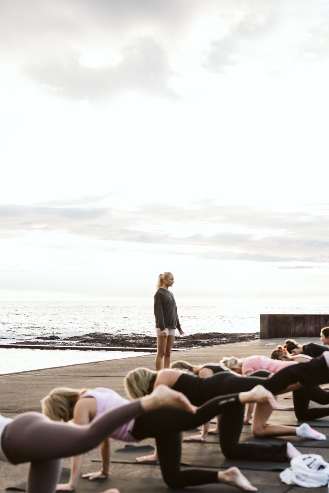 Fitness instructor leads outdoor yoga class on oceanfront platform at sunrise with cloudy sky.