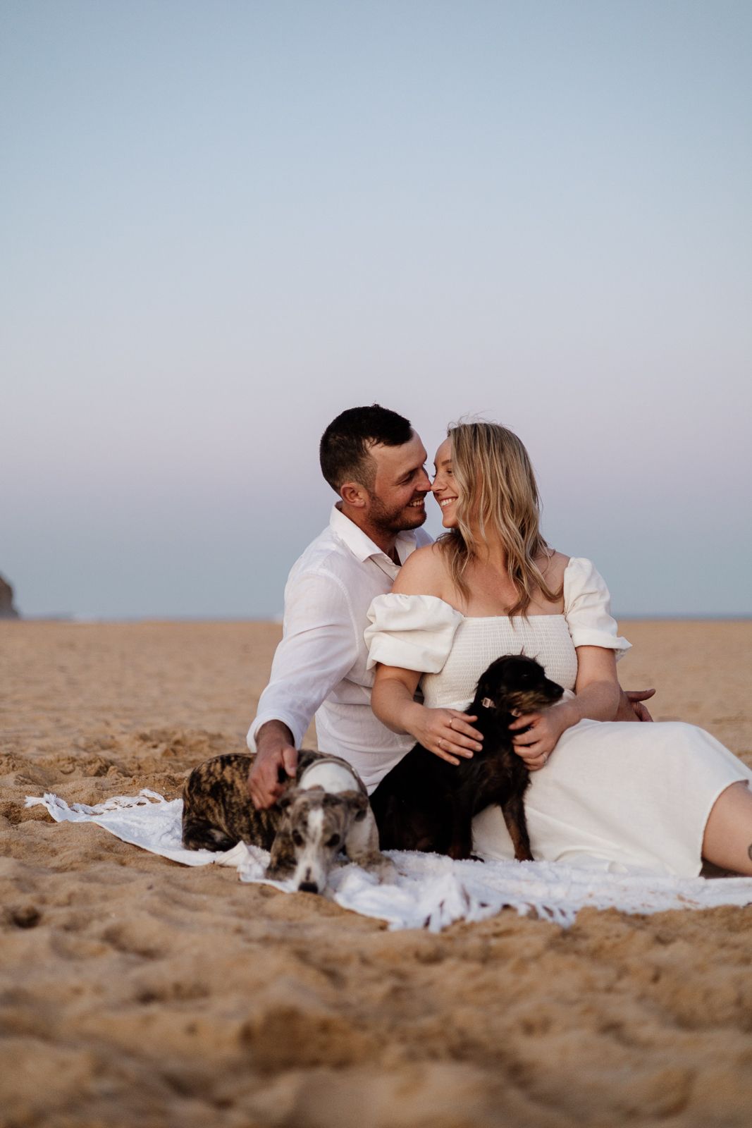 Happy couple sitting on beach blanket with two small dogs at sunset, smiling at each other.