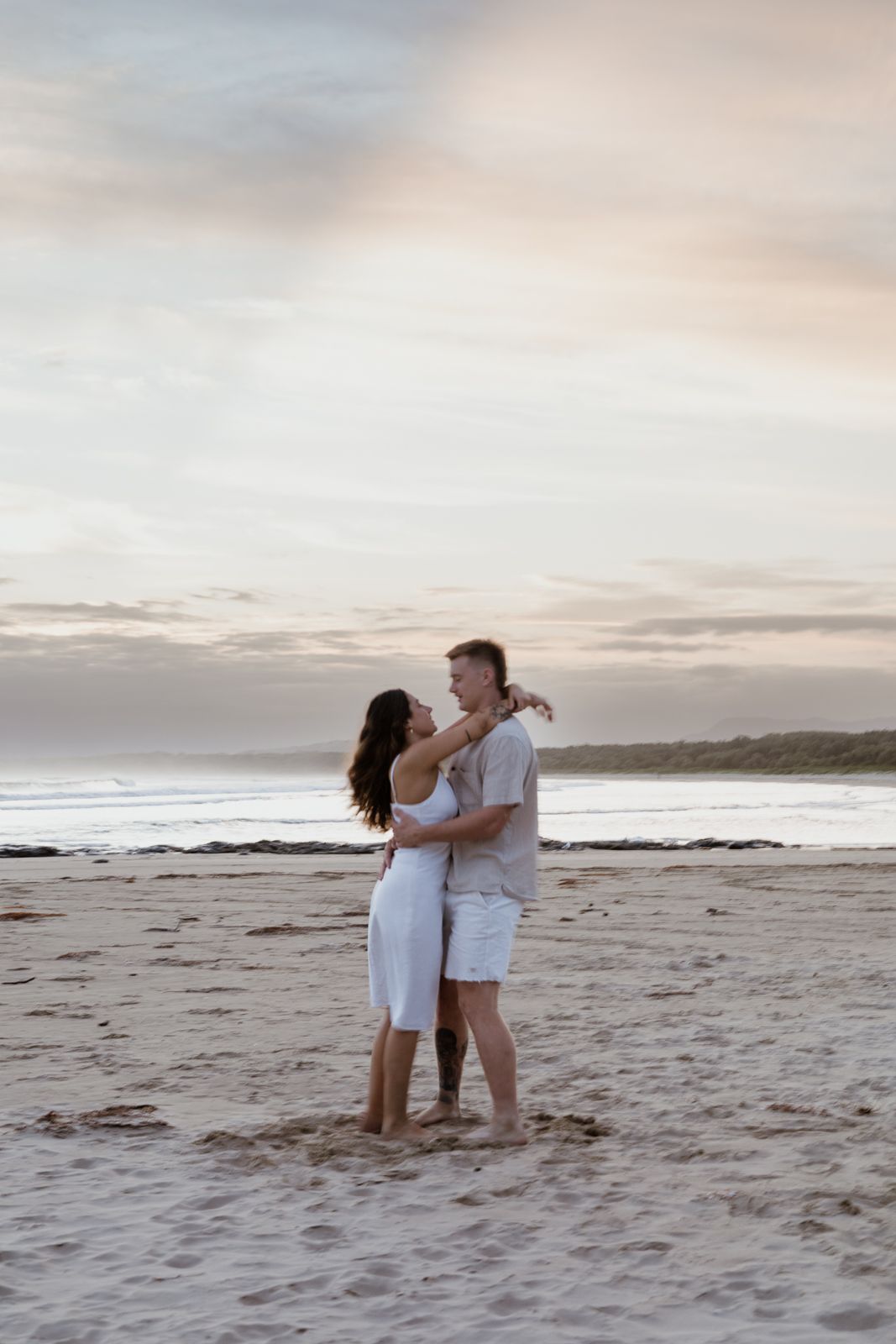 Couple dancing and laughing together on a sandy beach at sunset, dressed in white with ocean waves in the background.