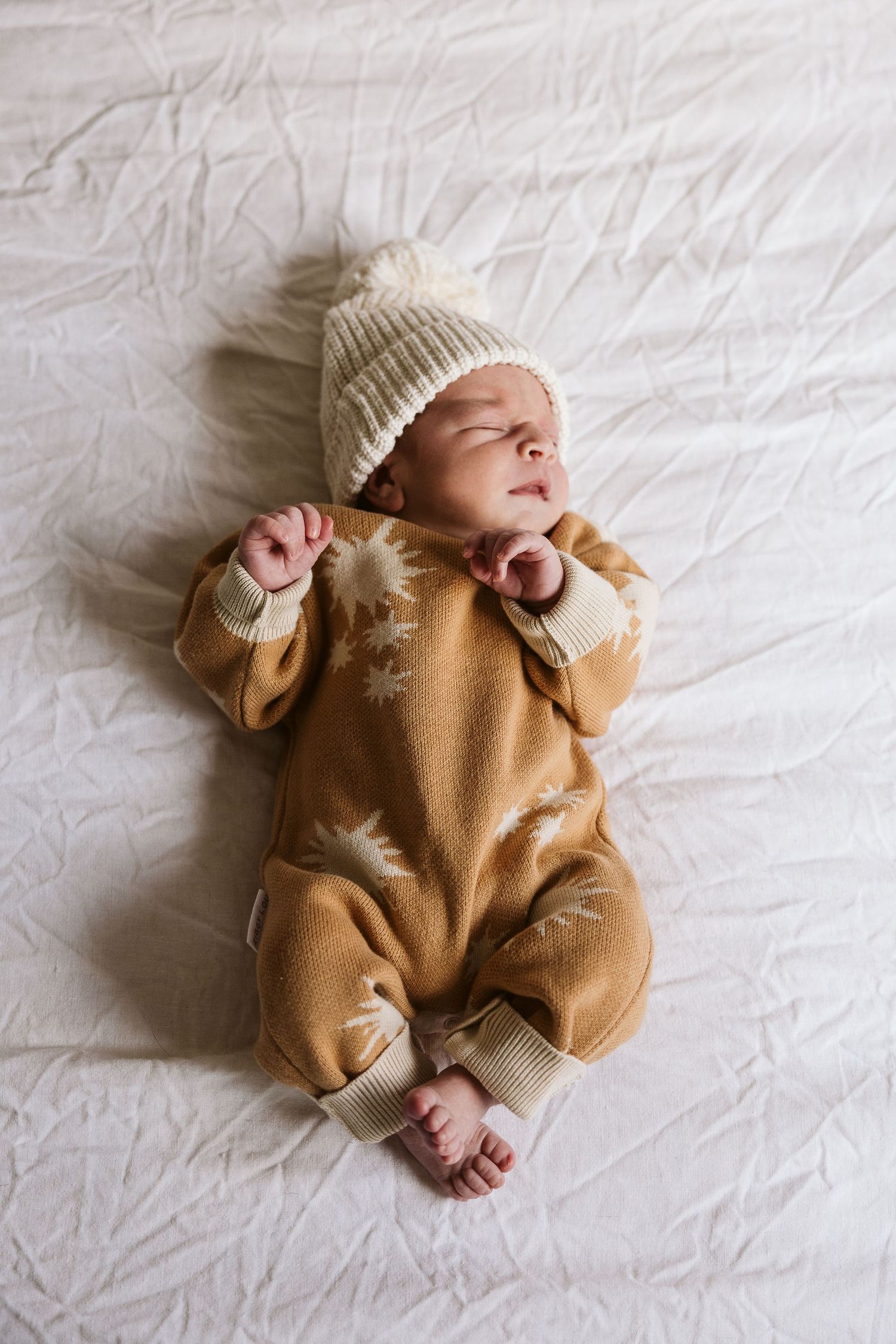 Sleeping newborn in tan star-print romper and cream pom-pom hat lying on white bed.