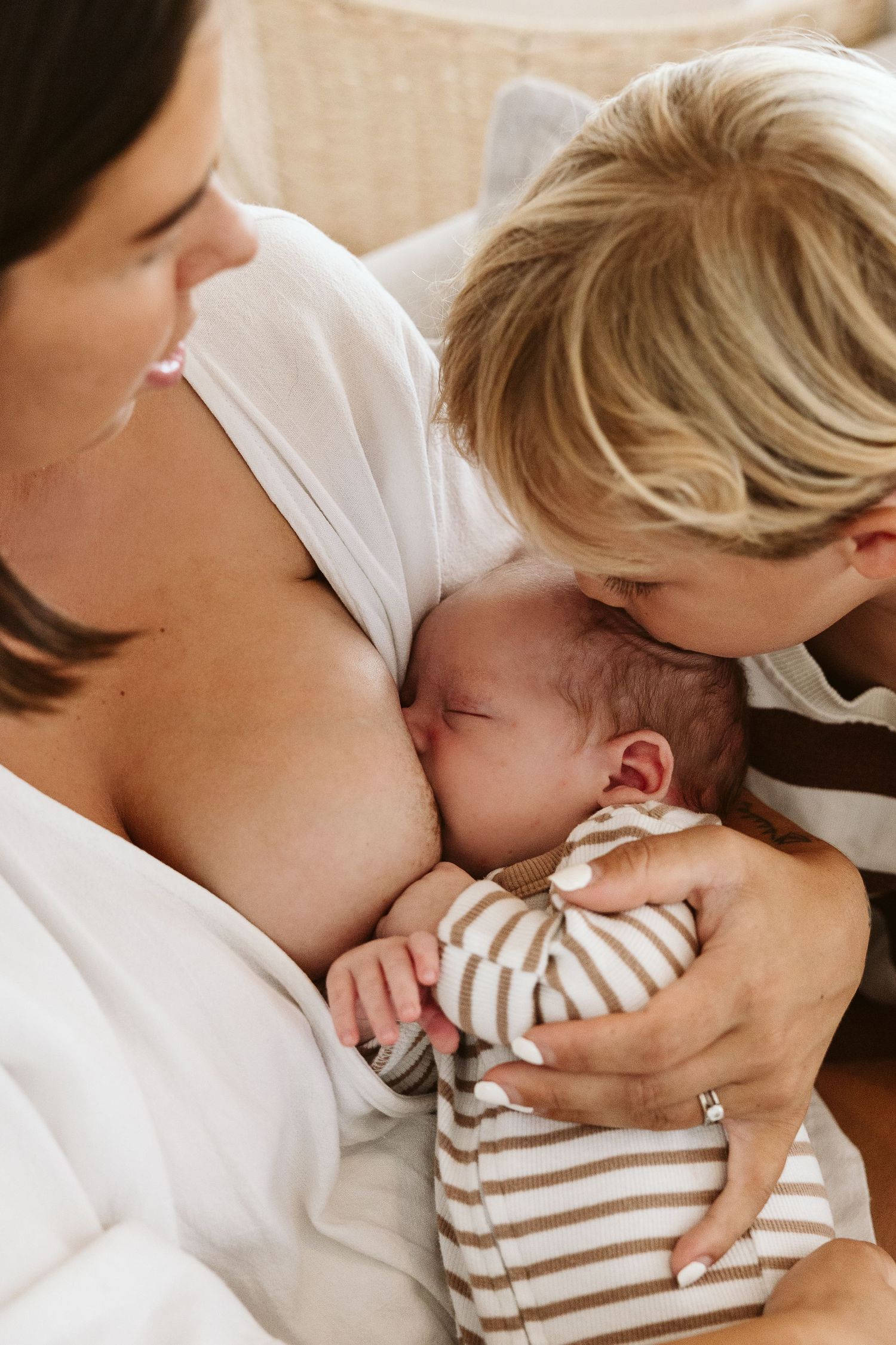 Mother breastfeeding newborn baby while older sibling gently kisses infant's head in warm home setting.