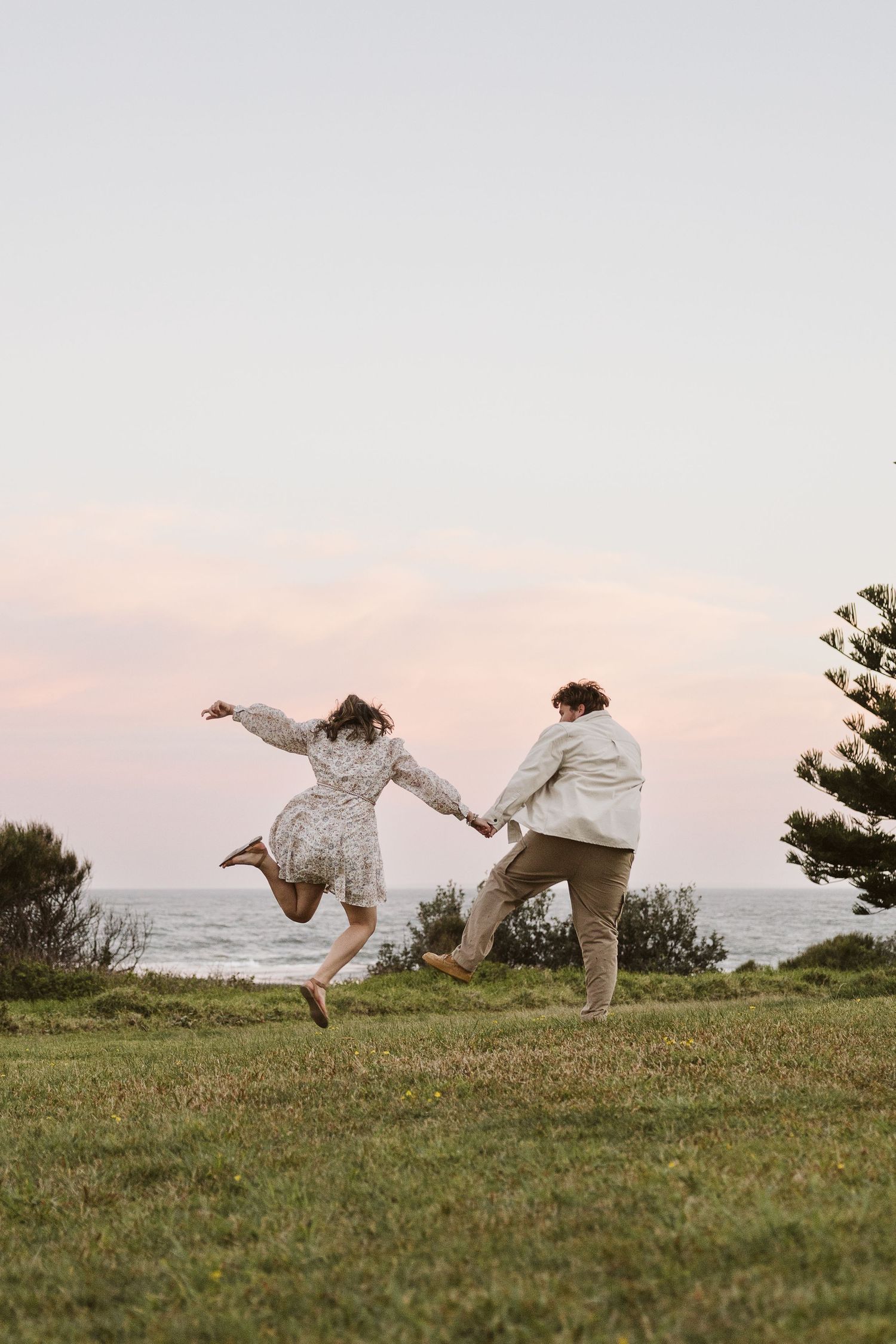 Joyful couple holding hands as woman leaps in the air on grassy cliff overlooking ocean at sunset.