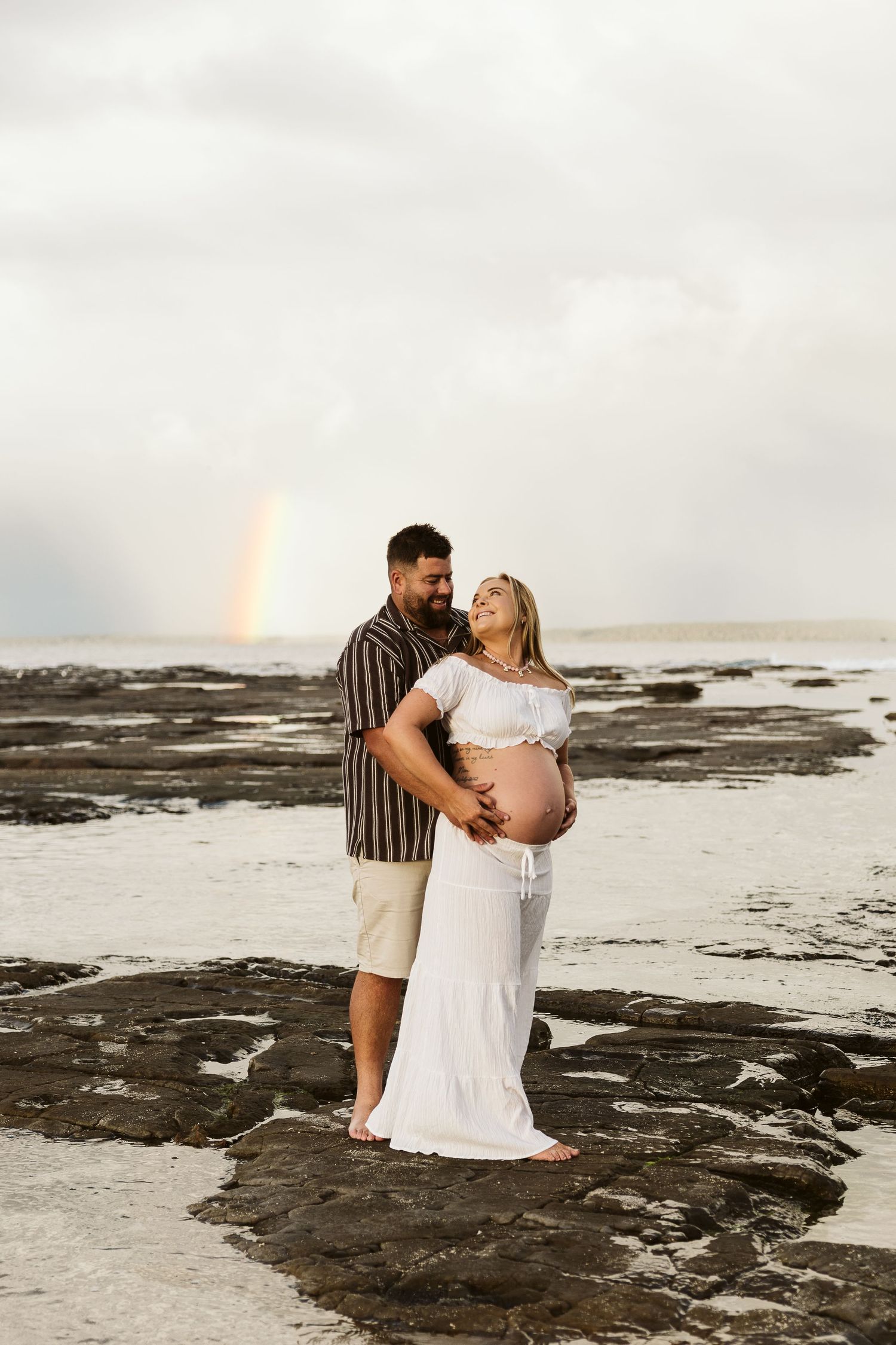 Couple shares tender moment on rocky beach, man embraces pregnant woman in white dress with rainbow behind.
