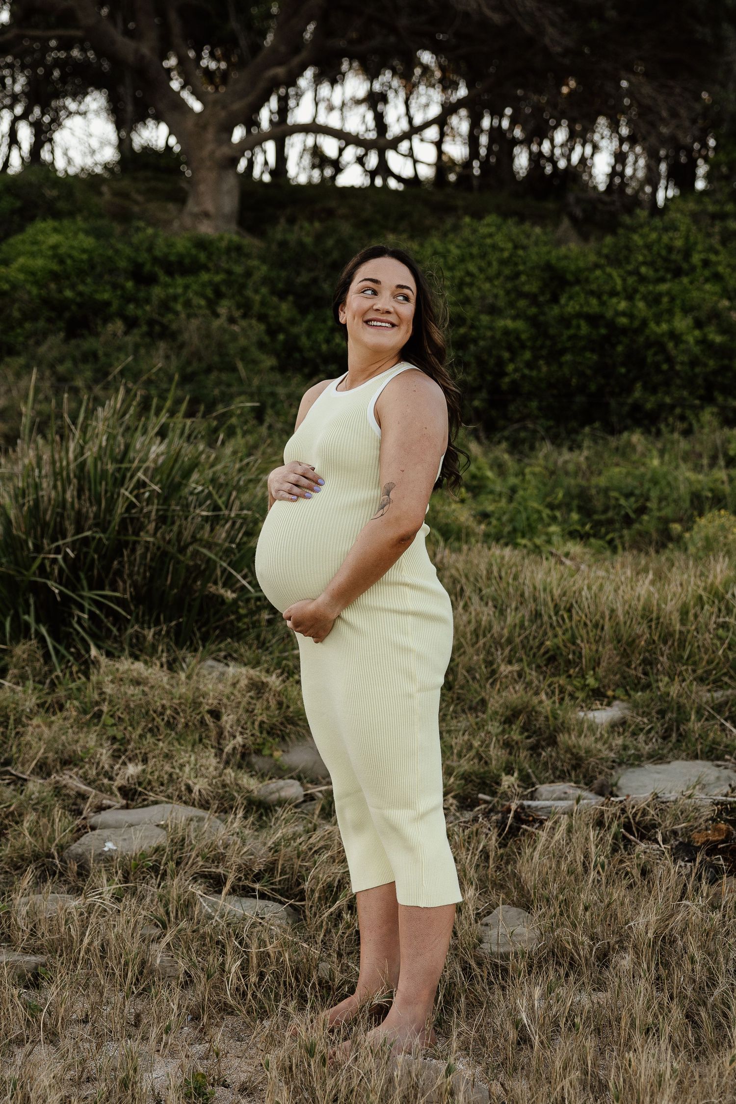 Smiling pregnant woman in yellow dress cradles belly standing in natural greenery.