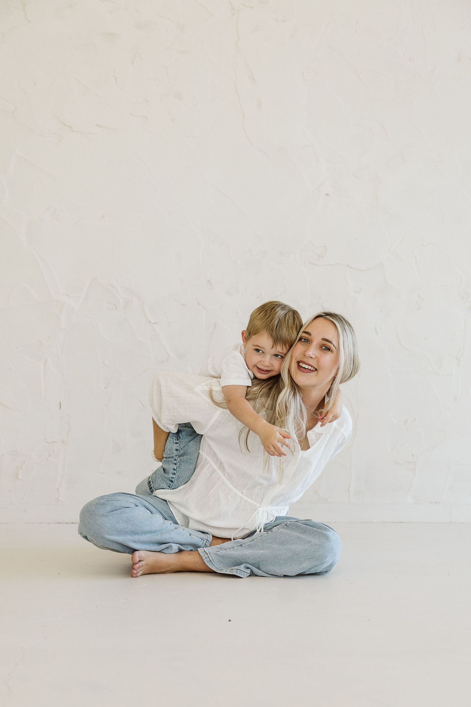 A smiling blonde mother sits cross-legged while her young son hugs her from behind against a white wall.