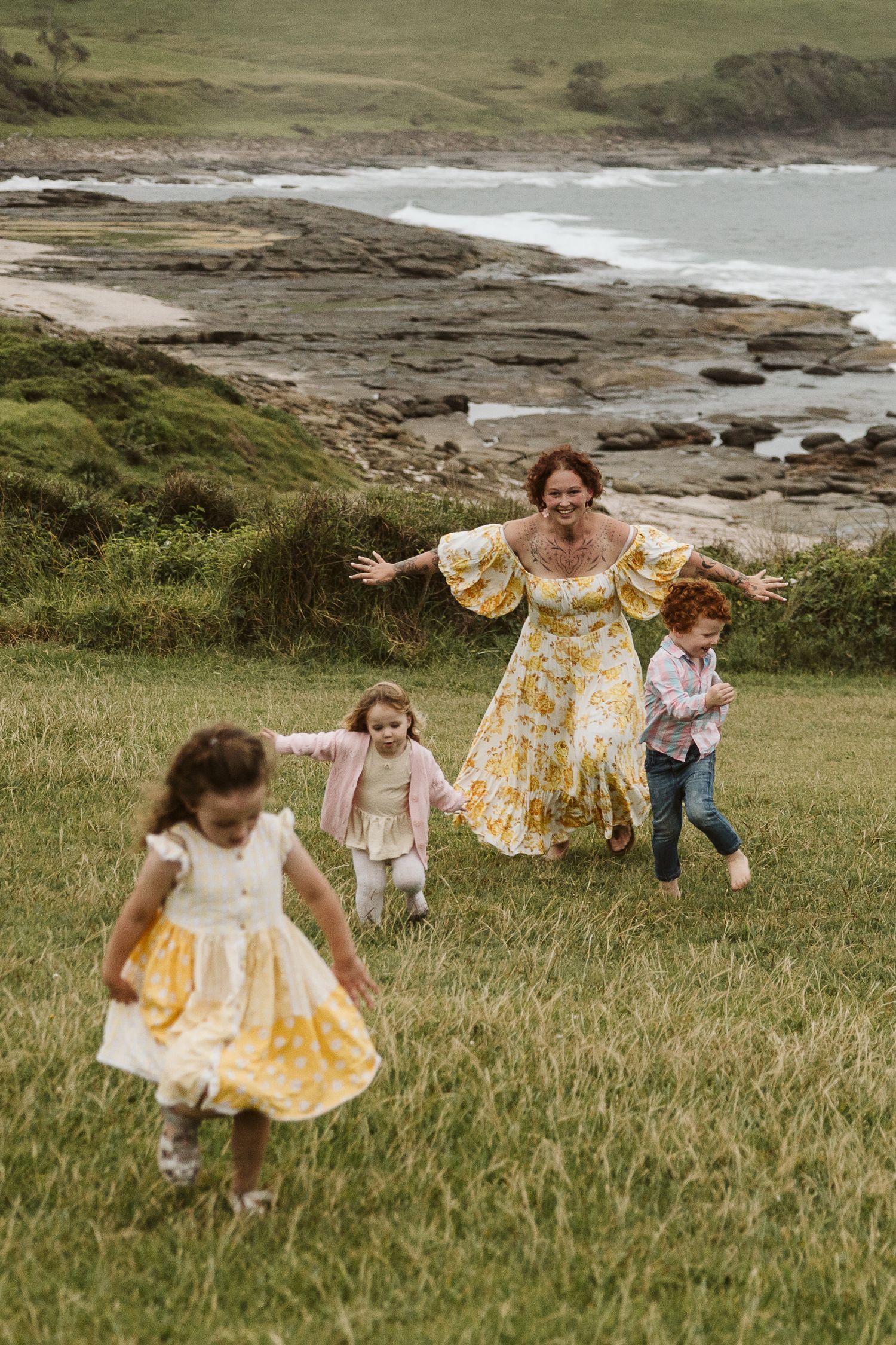 Joyful mother in floral dress running with three children on a grassy coastal hillside.