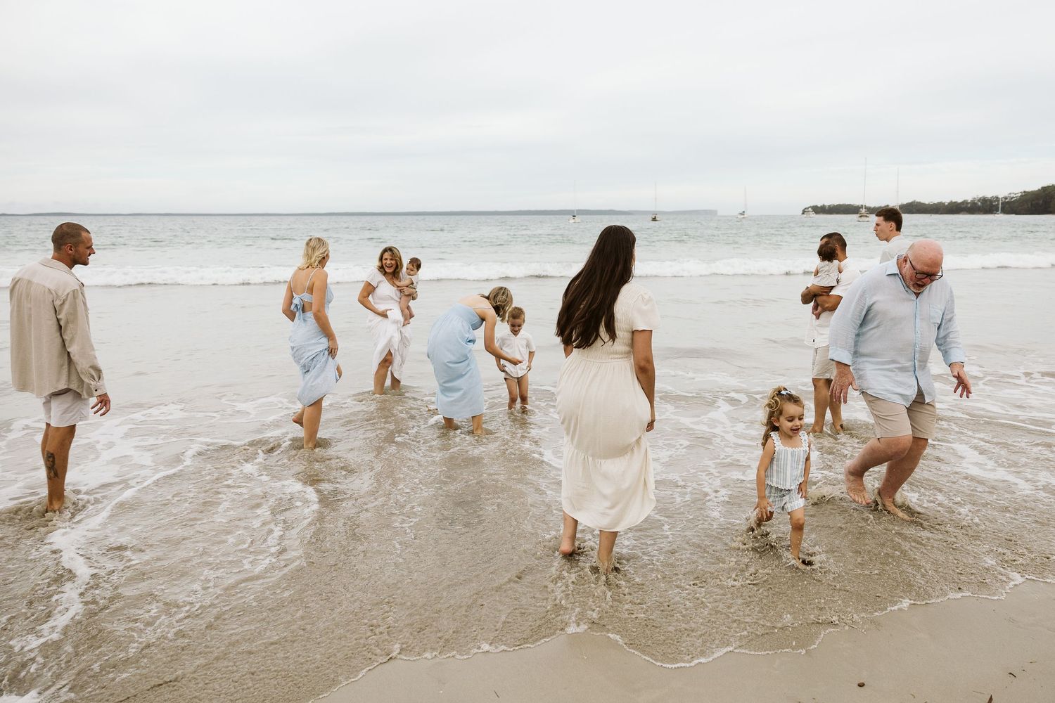 Large family gathering on a sandy beach, wading in ocean waves, dressed in coordinated white and blue outfits.