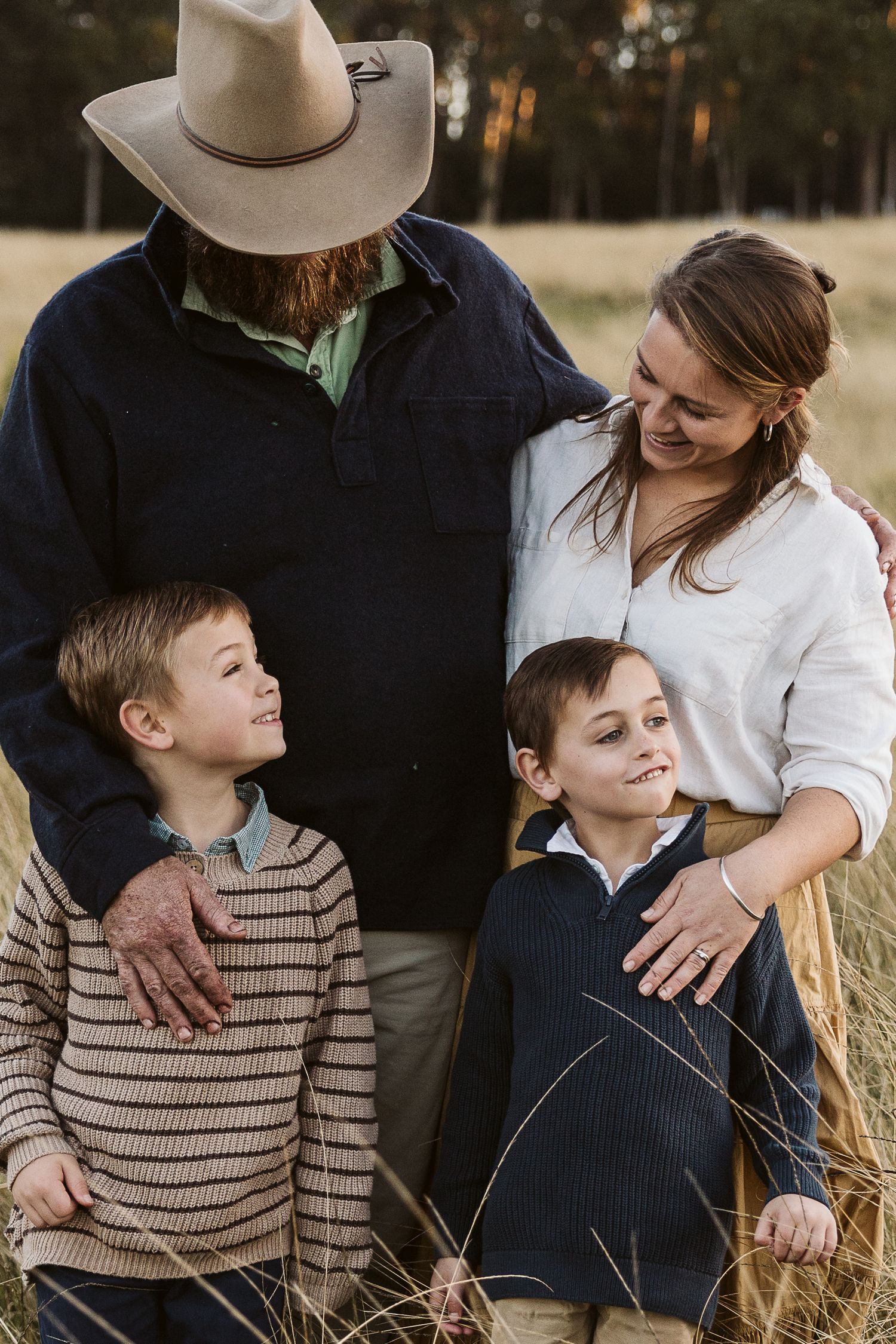 A family of four poses warmly in a golden grass field, with a bearded man in a cowboy hat and two young boys.