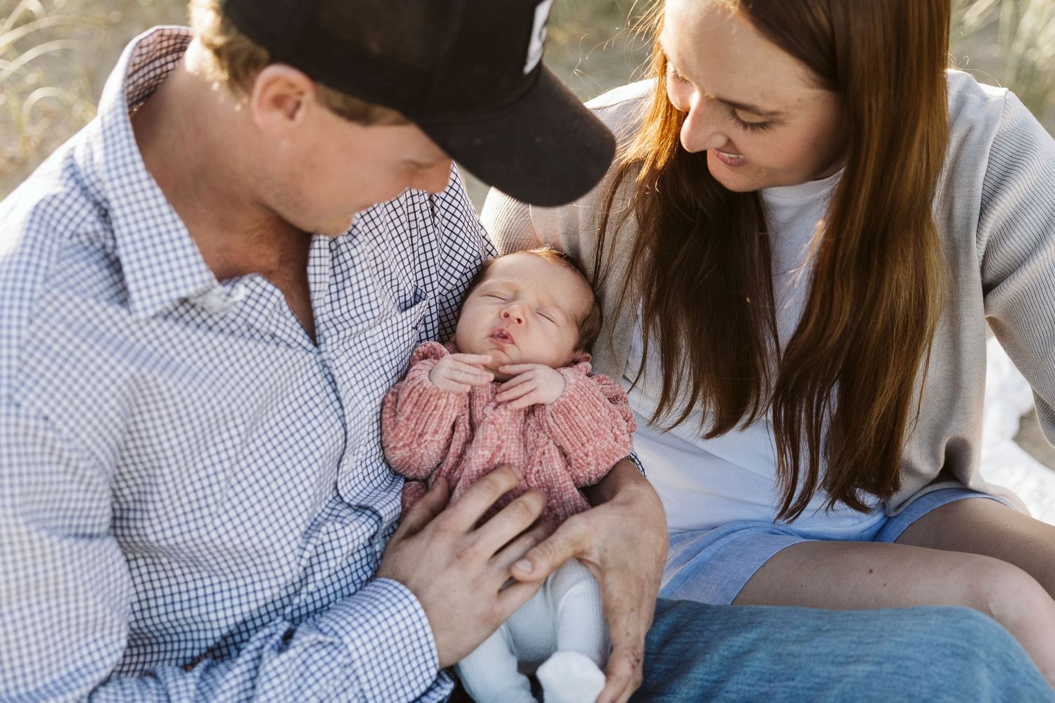 New parents sitting outdoors together admiring their sleeping newborn in pink outfit.