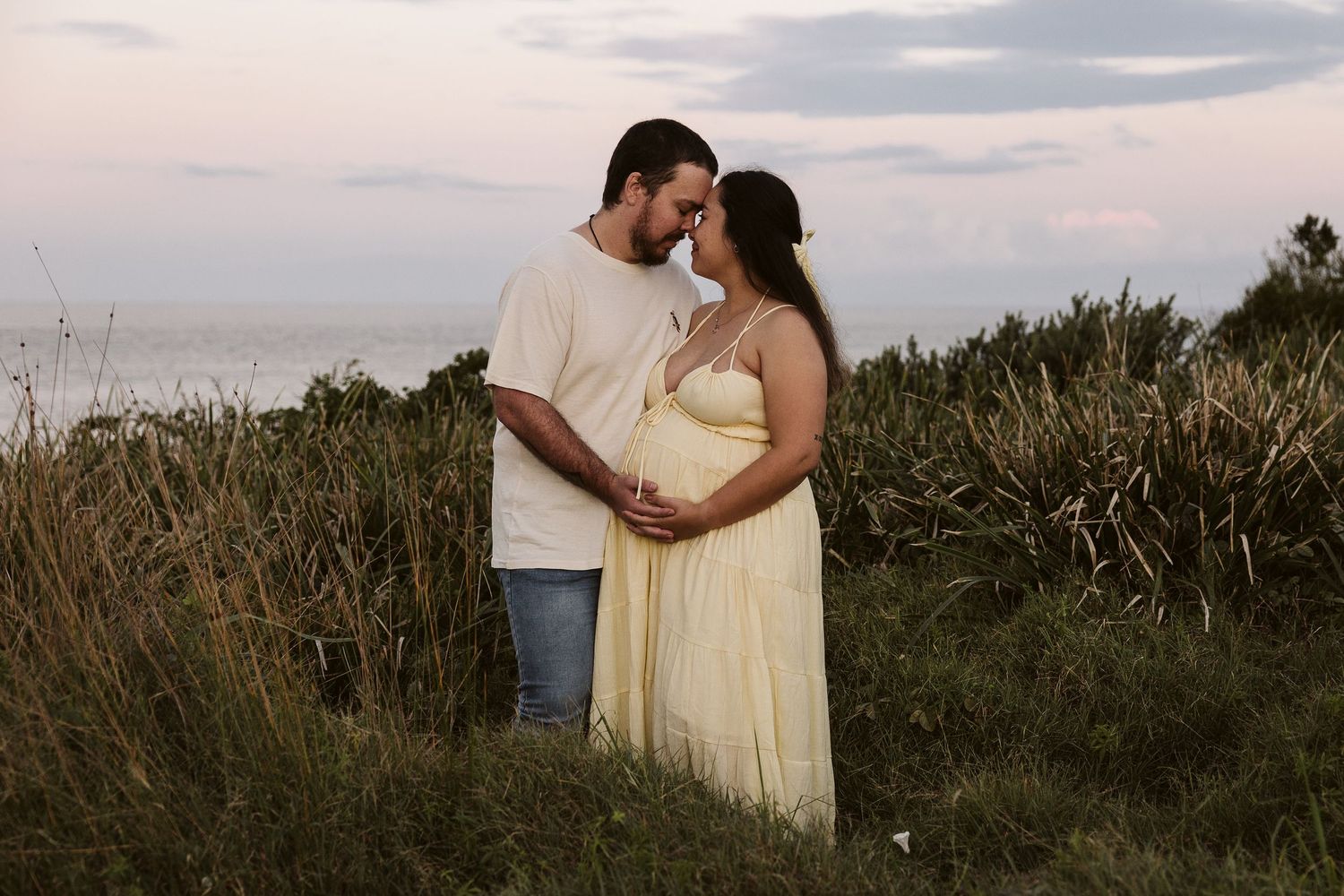 Expecting couple sharing a tender moment in coastal grass field at dusk.