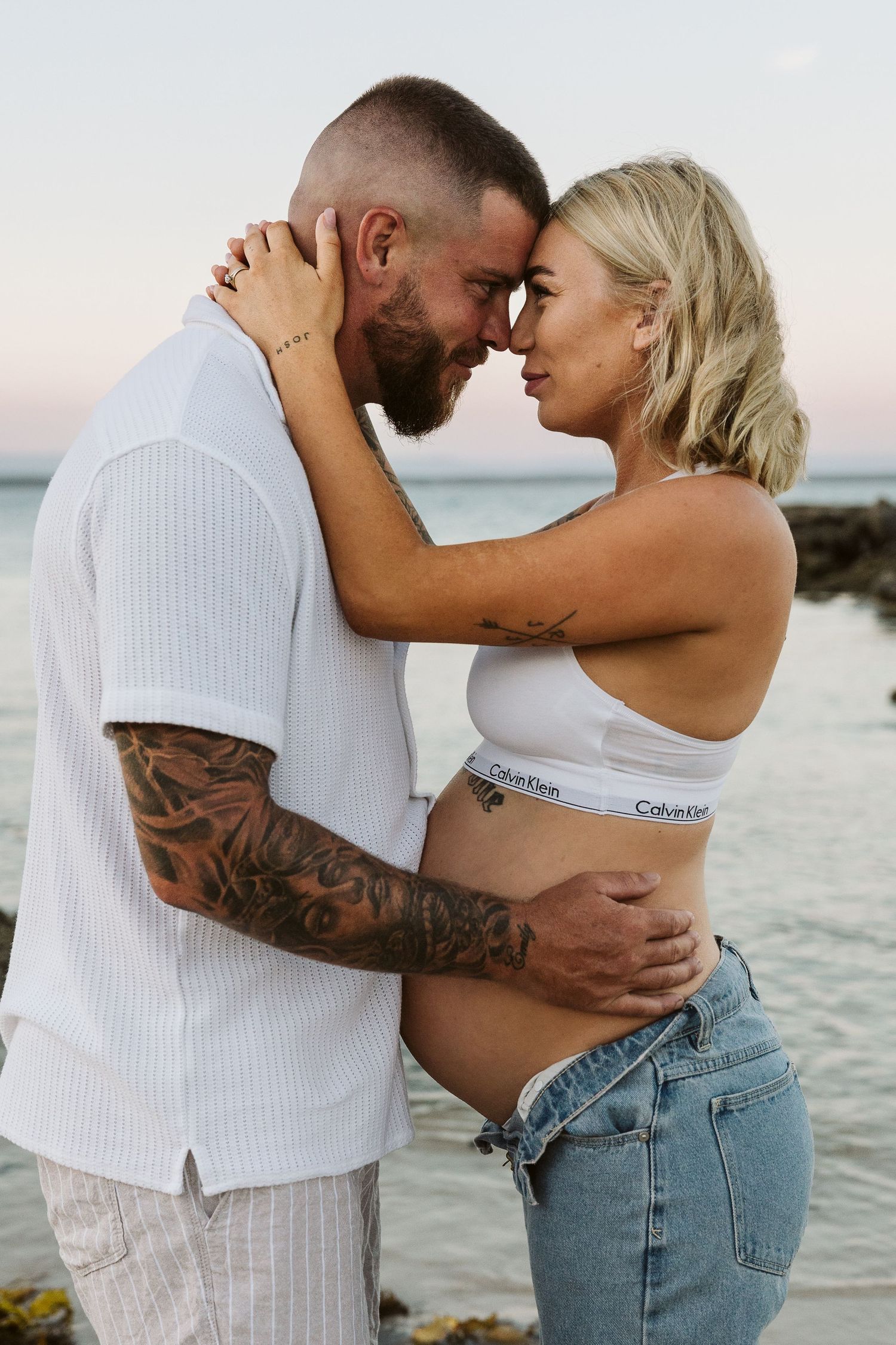 Tattooed couple touching foreheads intimately on rocky beach, man embracing pregnant woman's baby bump.