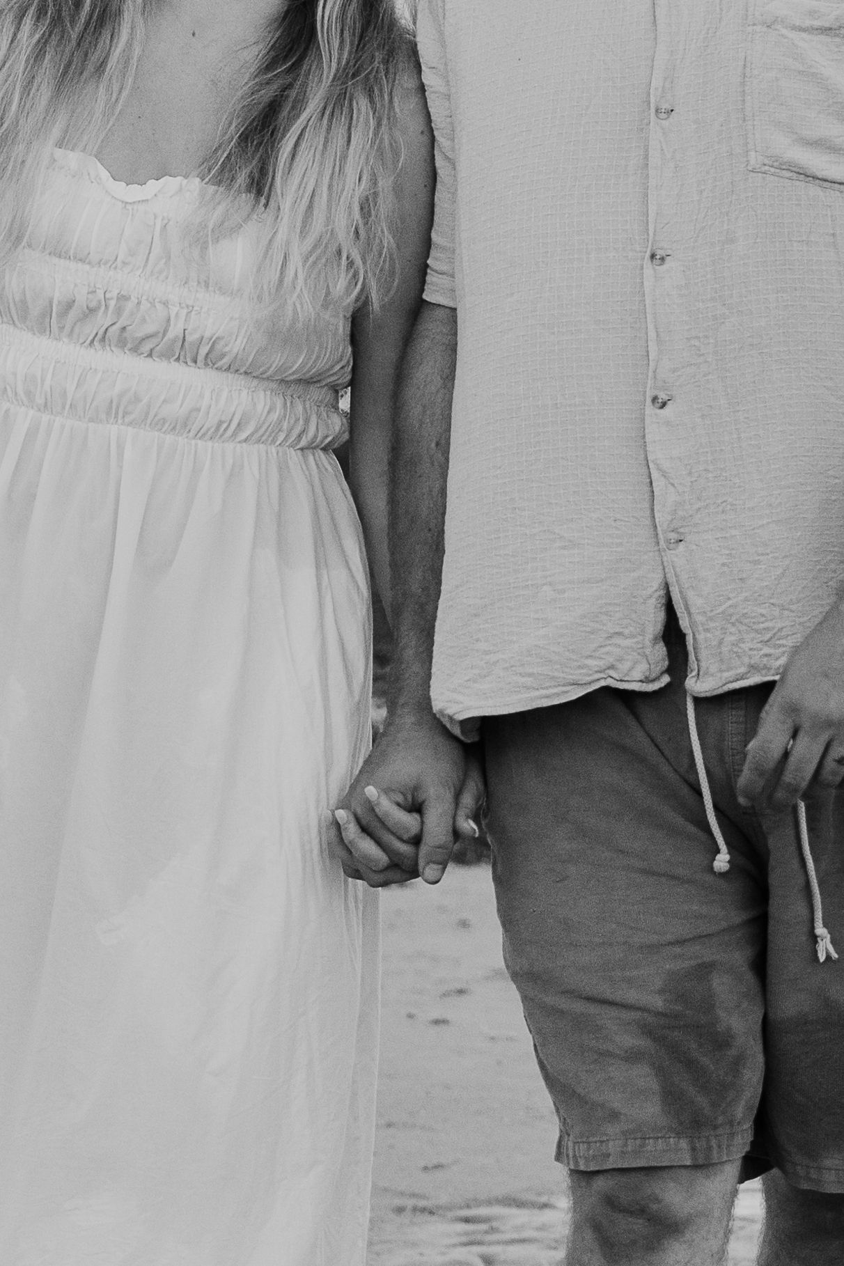 Black and white photo of a couple holding hands, woman in white dress and man in casual shorts and button-up shirt.