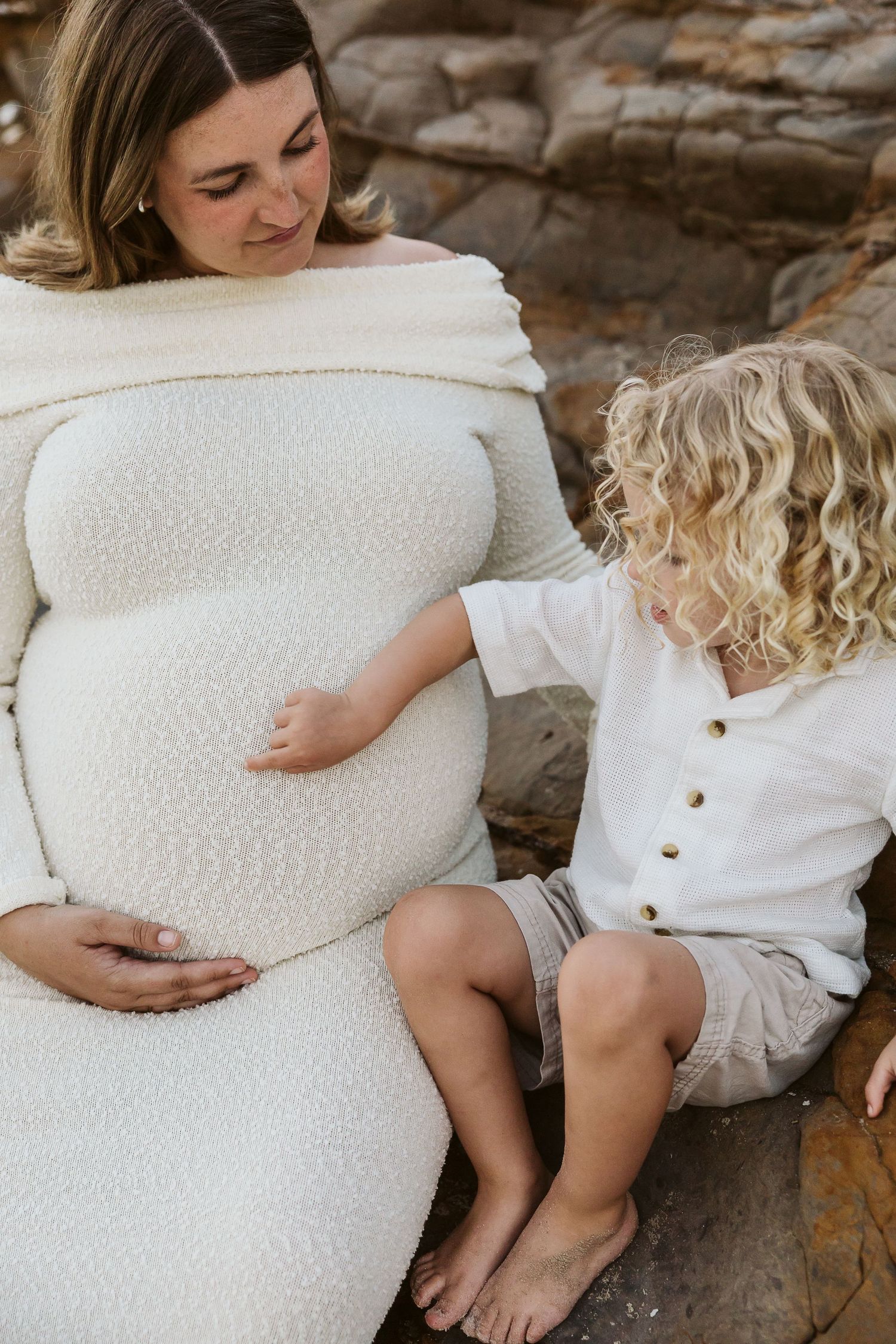 Pregnant woman in cream off-shoulder dress sits on rocks with curly-haired toddler touching her baby bump.