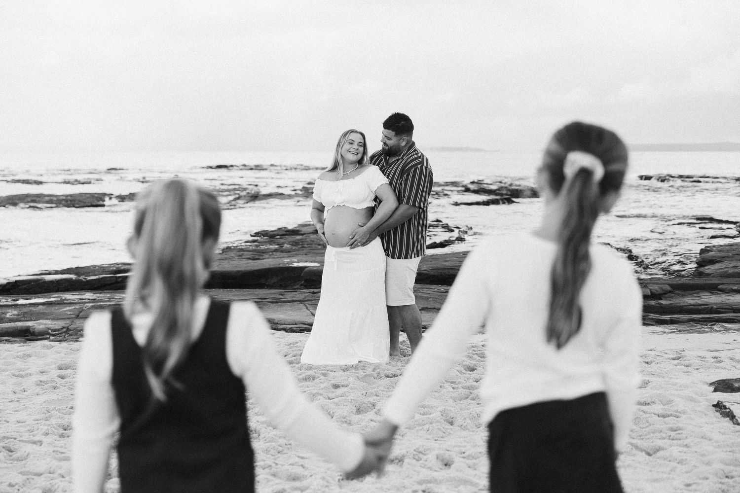 Black and white beach photo of couple holding hands with two girls in foreground.