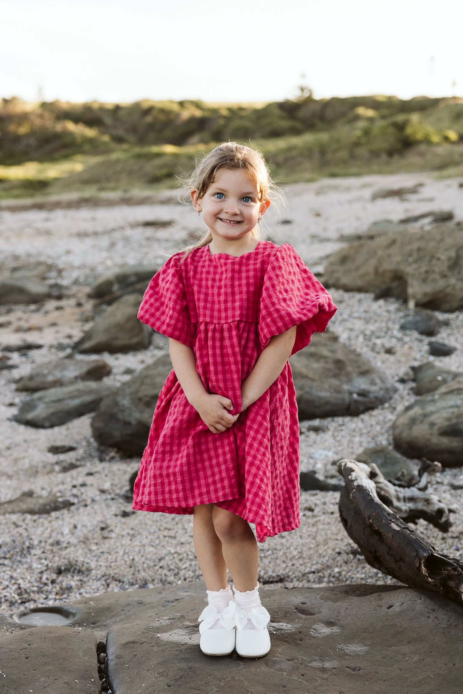 A smiling little girl in a red gingham dress with puff sleeves stands on rocks at a pebble beach.