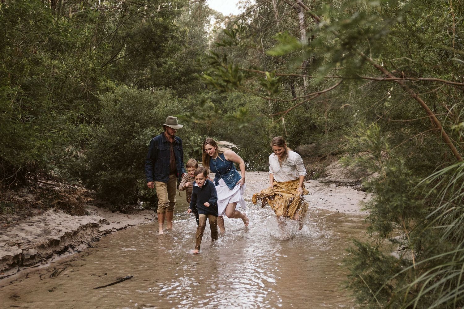 Family splashing through a shallow creek in a lush Australian forest, laughing and playing together outdoors.