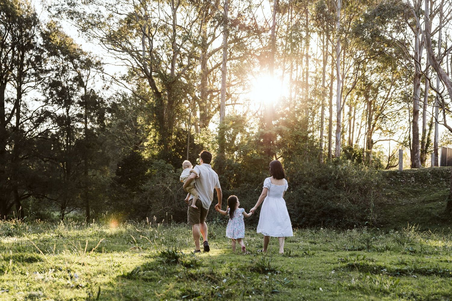 Family walking hand-in-hand through sunlit forest clearing at golden hour, parents carrying young child.