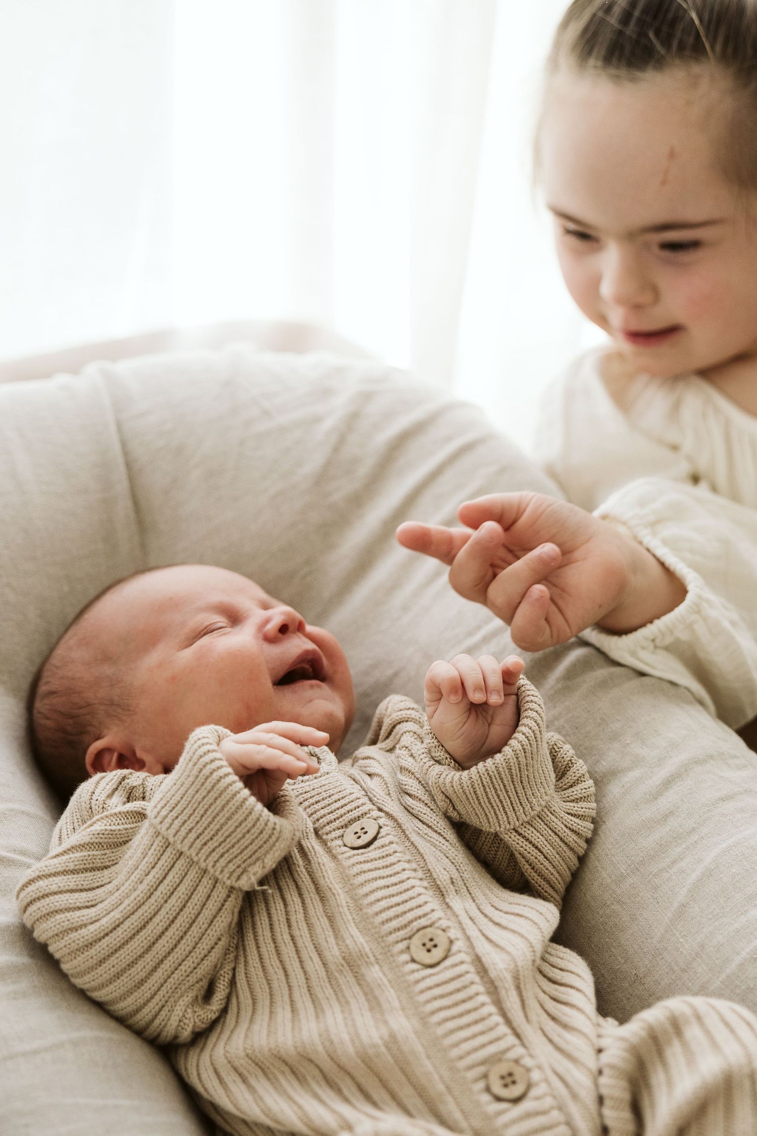 Young girl gently pointing at a laughing newborn baby resting in a soft white lounger indoors.