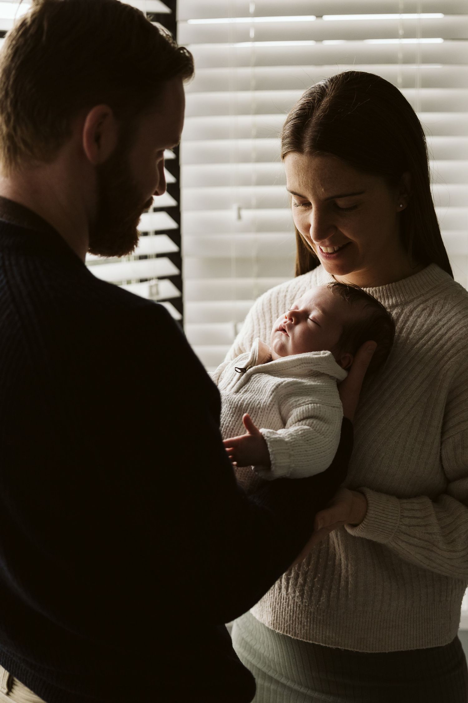Couple smiles down at their newborn baby by a window with soft light.