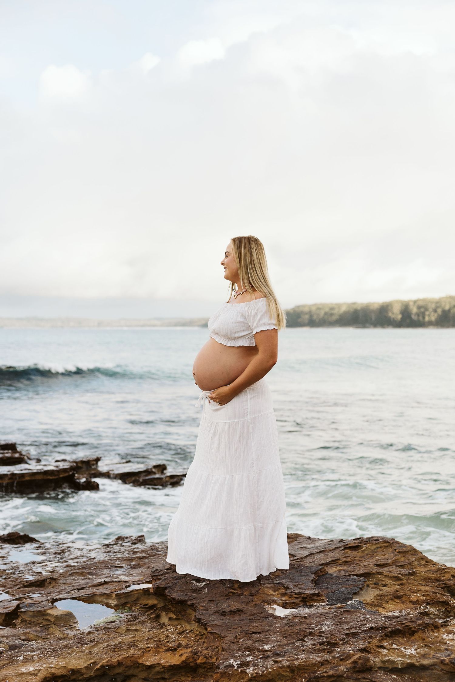 Pregnant woman in white off-shoulder dress stands on ocean rocks at dusk.