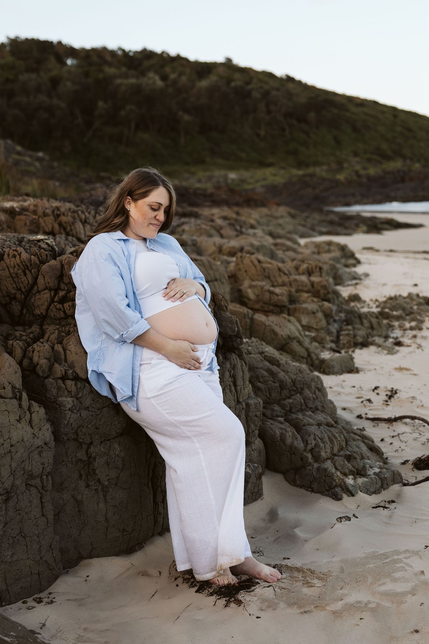 Pregnant woman in blue shirt and white skirt leaning against coastal rocks on sandy beach, cradling bump.