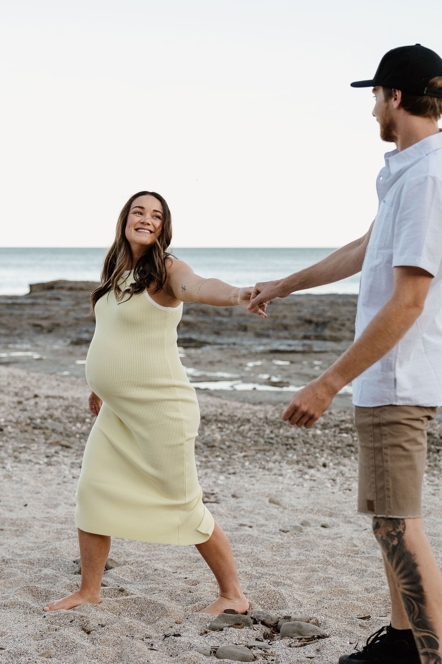 Smiling pregnant woman in yellow dress holding hands with tattooed man on rocky beach at dusk.