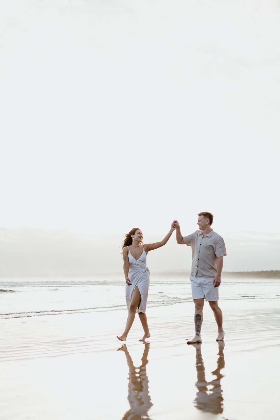 Couple dancing and holding hands on a reflective wet beach shore under a bright overcast sky.