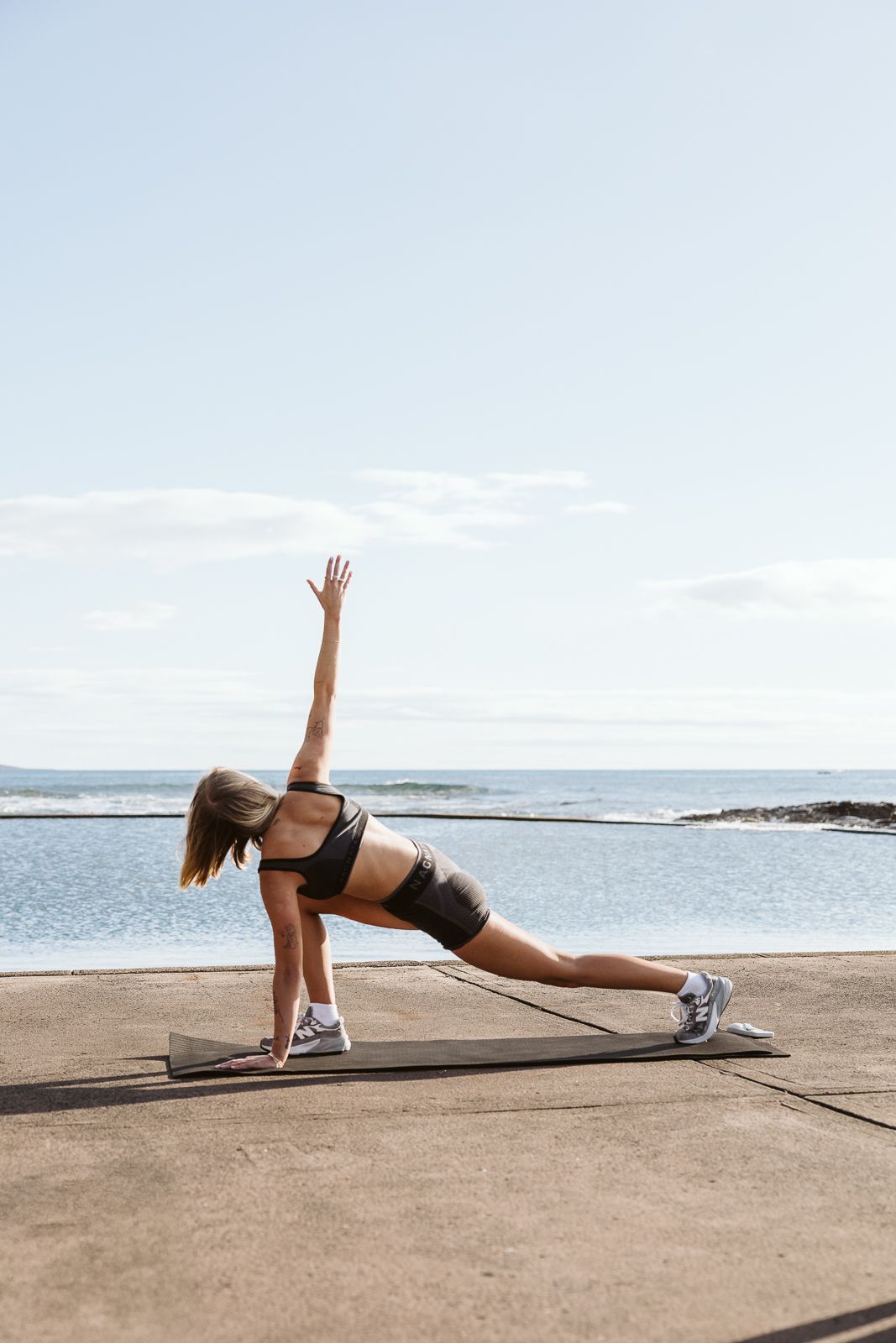 Female athlete performs side plank variation in sportswear on outdoor mat by ocean.