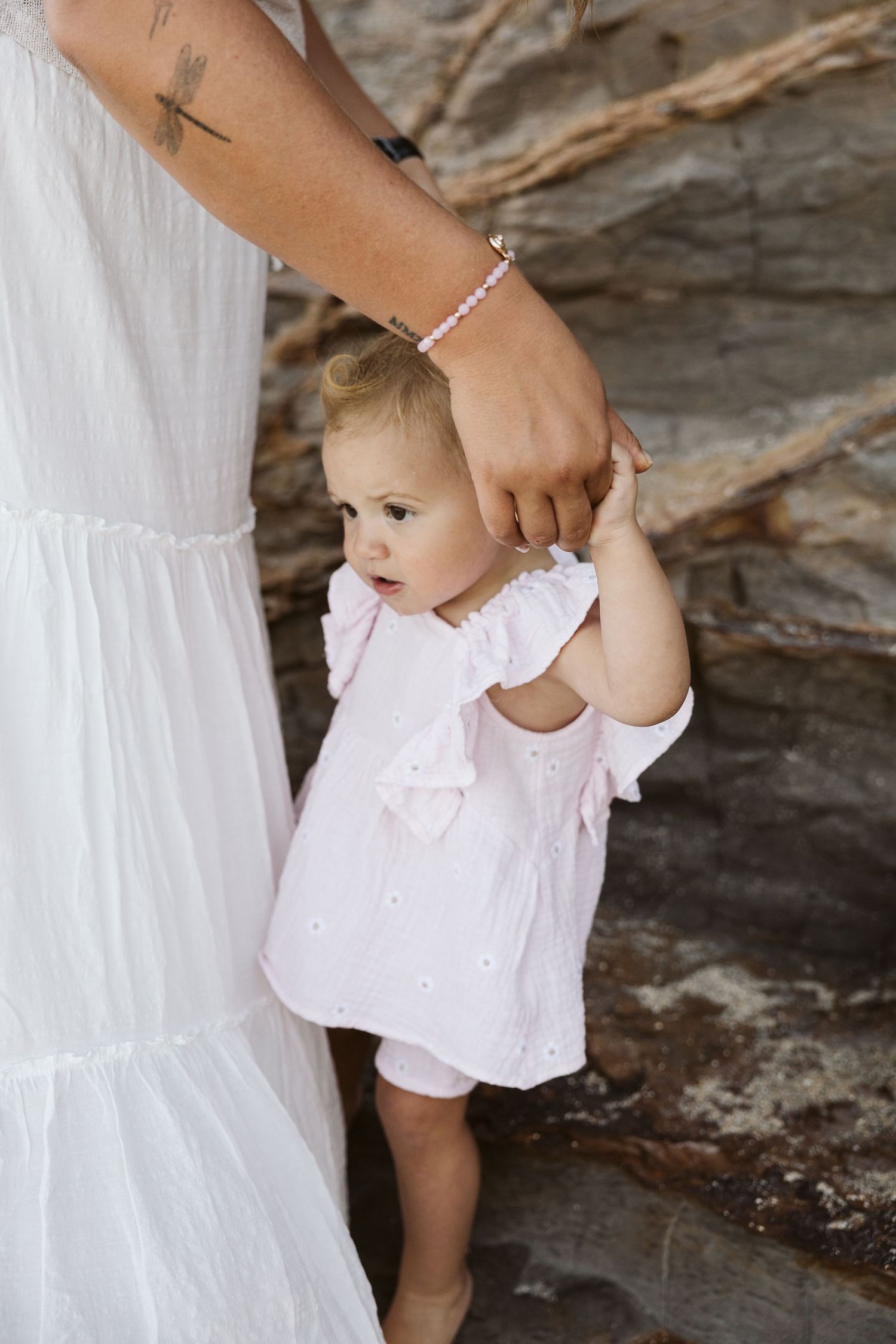 A toddler in a pink dress holds her mother's hand while learning to walk near rocky terrain outdoors.