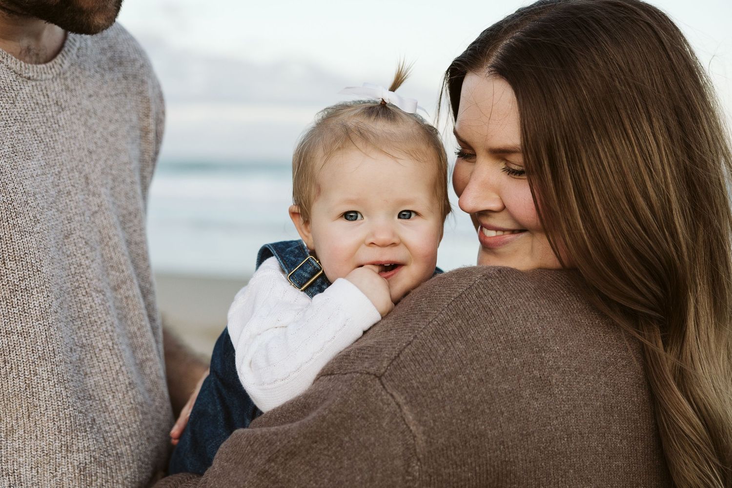 A smiling mother holds her happy baby on a beach, with a man in a gray sweater beside them.