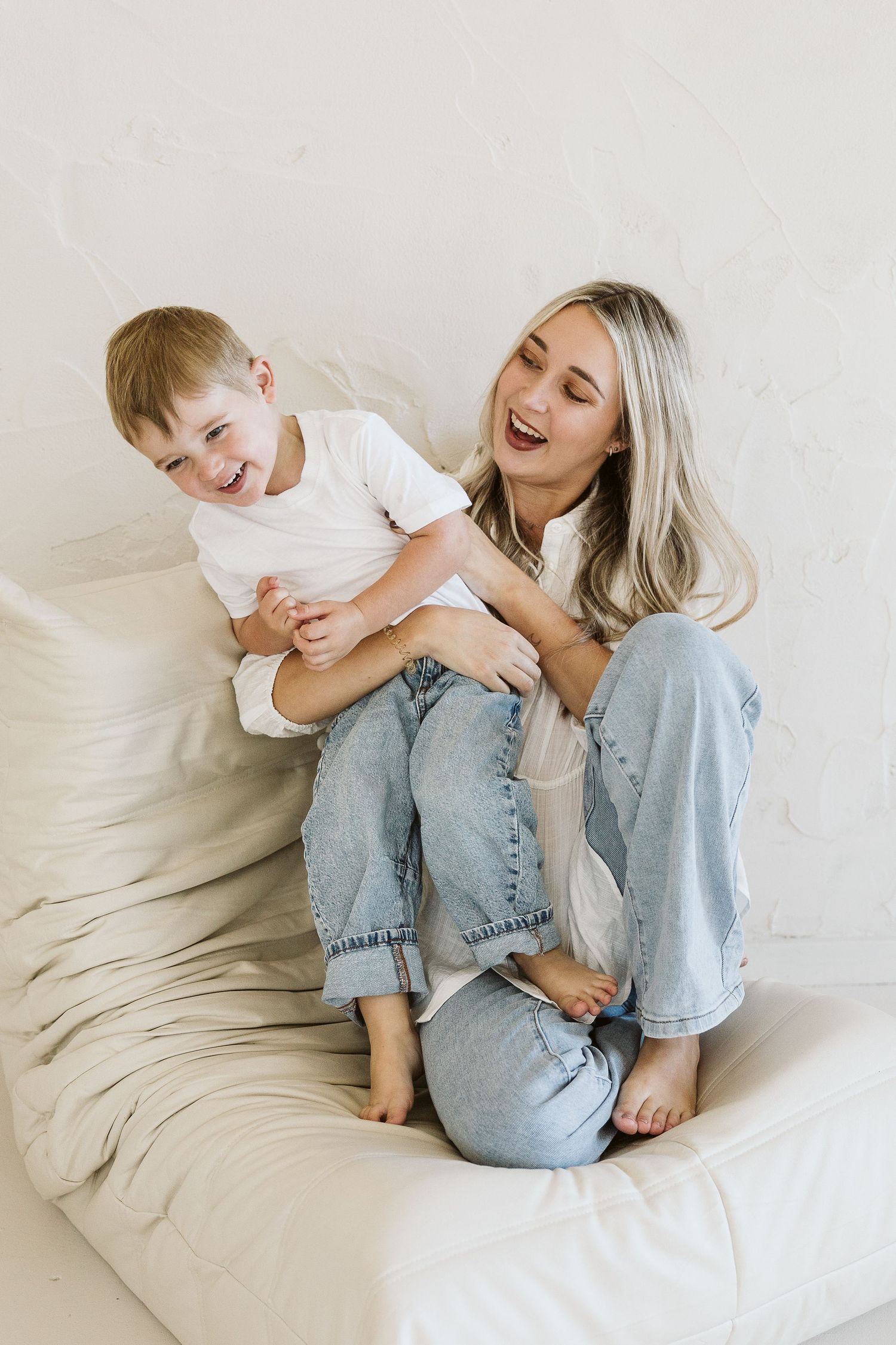 A laughing blonde mother in jeans playfully holds her giggling toddler son on a cream sofa.
