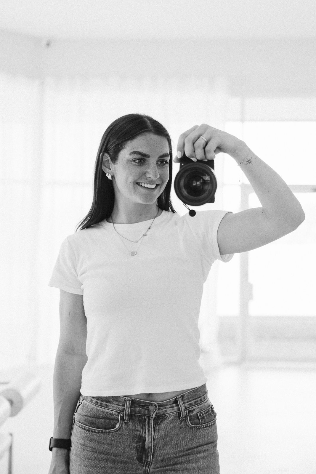 Smiling brunette woman holding a camera in a bright white studio in black and white photo.