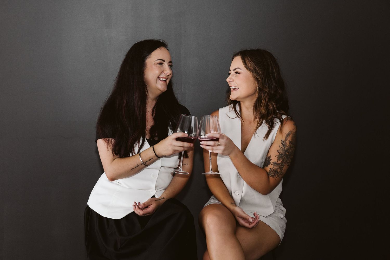 Two women smiling and clinking red wine glasses against dark background.