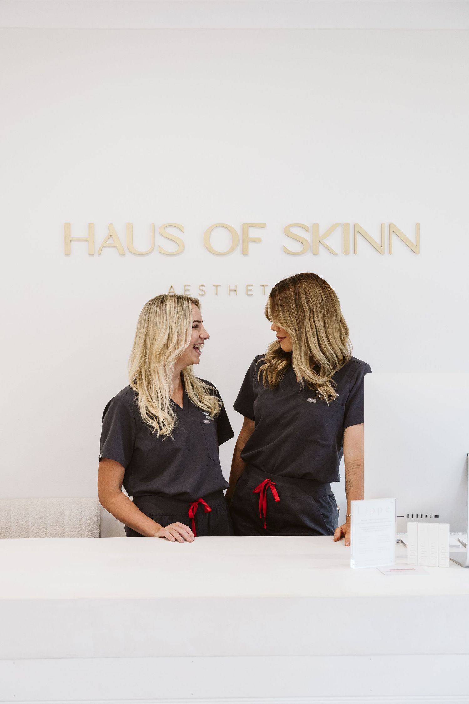 Two blonde female aestheticians in black scrubs smiling at each other behind reception desk at Haus of Skinn Aesthetics.
