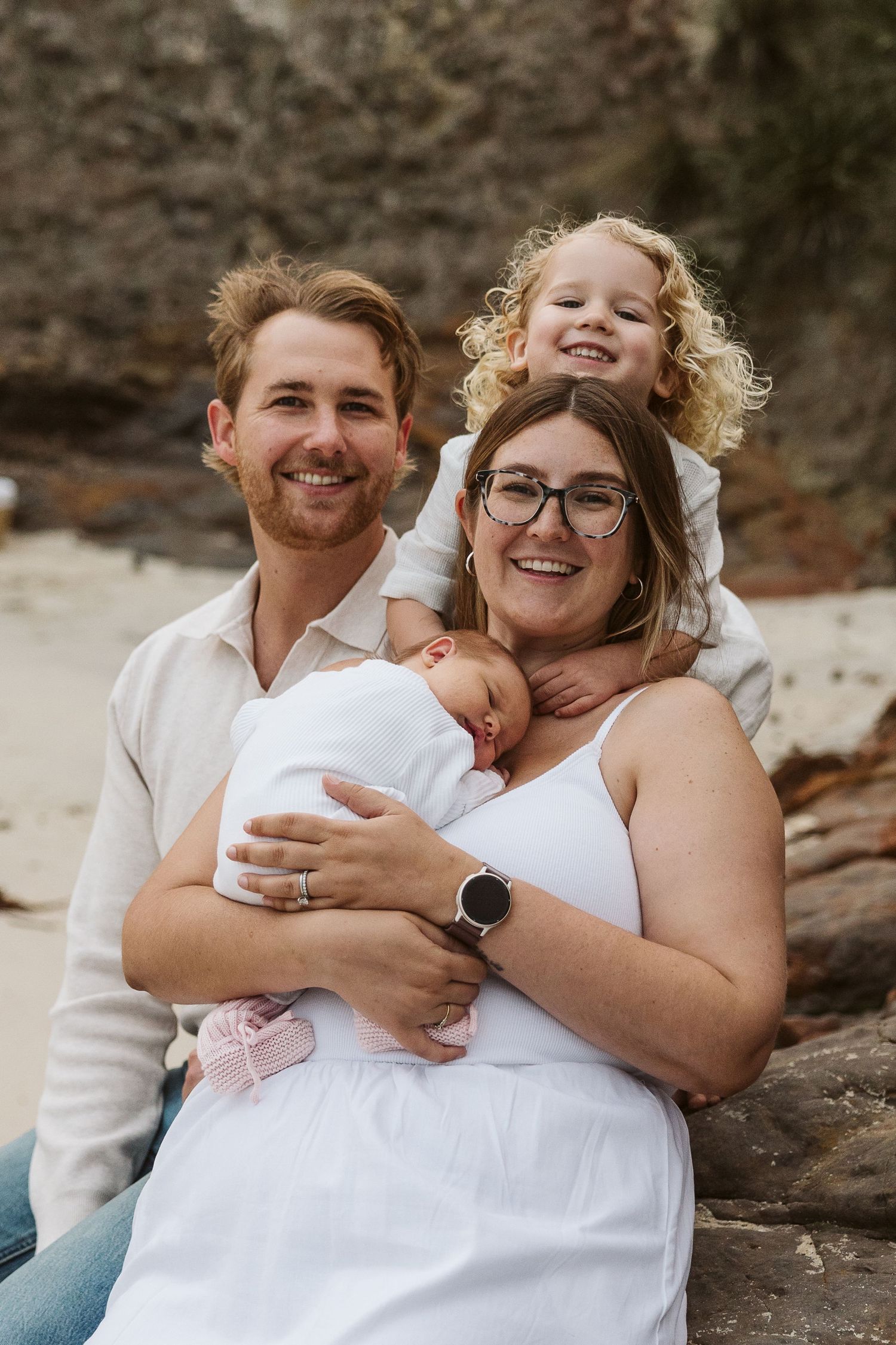 Smiling family of four with newborn baby and curly-haired child on beach rocks.