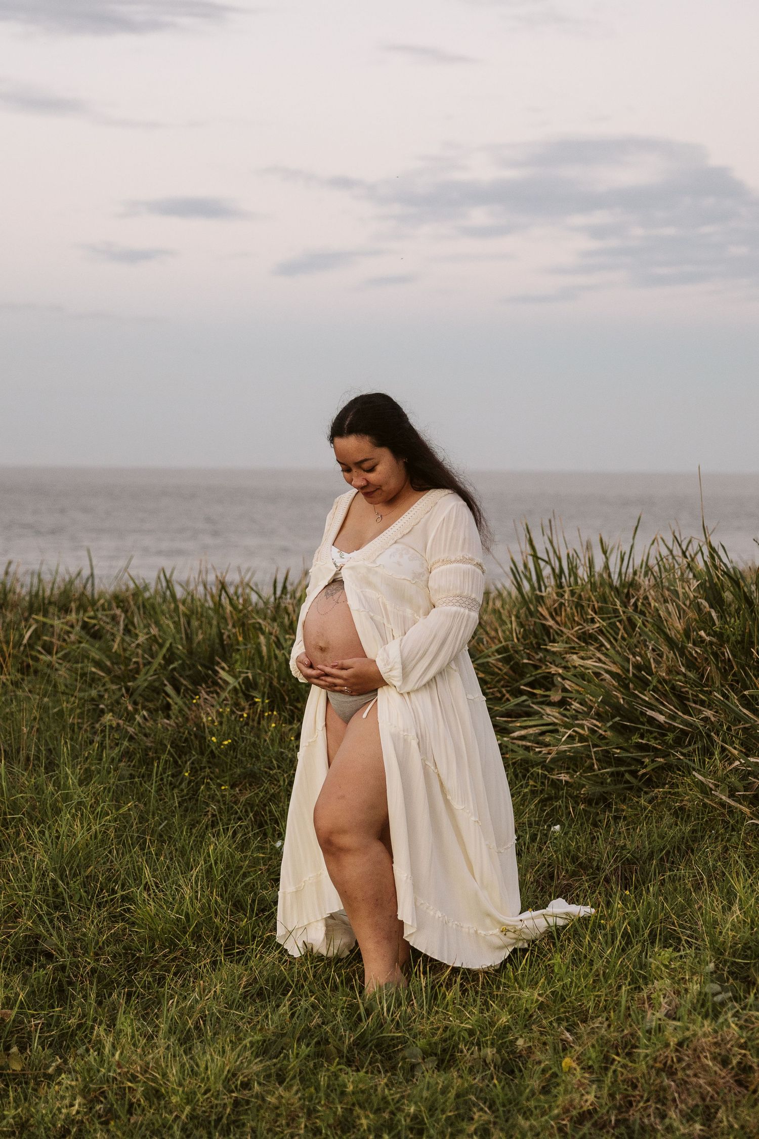 Pregnant woman in flowing white open robe cradling bump by ocean at dusk.