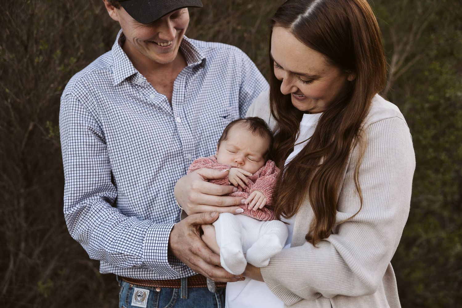 Smiling new parents holding sleeping newborn baby in pink outfit in nature setting.