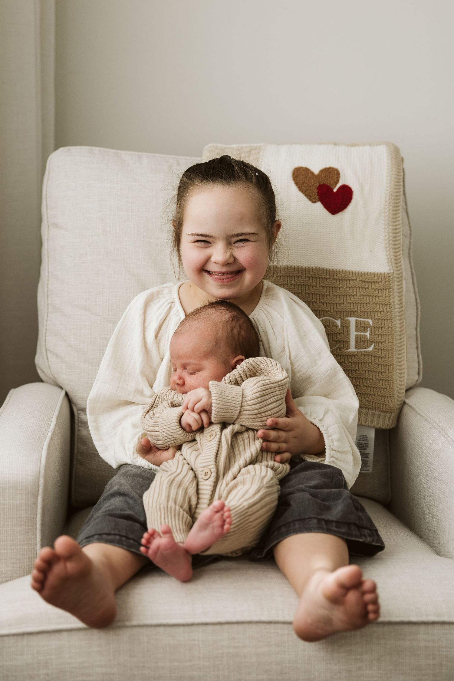Young girl with Down syndrome smiling while holding newborn sibling in a white armchair.