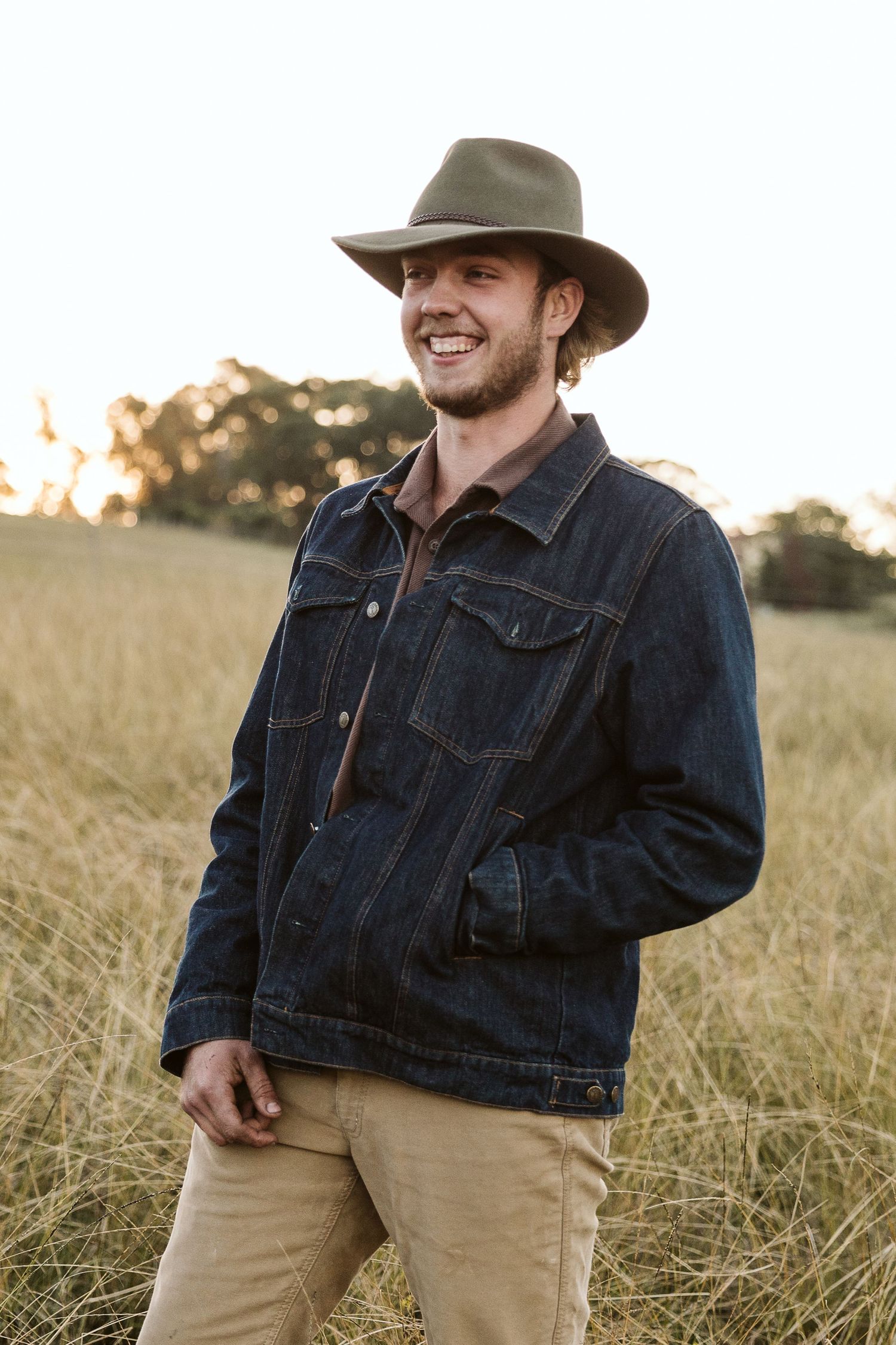 A smiling young man wearing a wide-brim hat and denim jacket stands in a golden grass field at sunset.