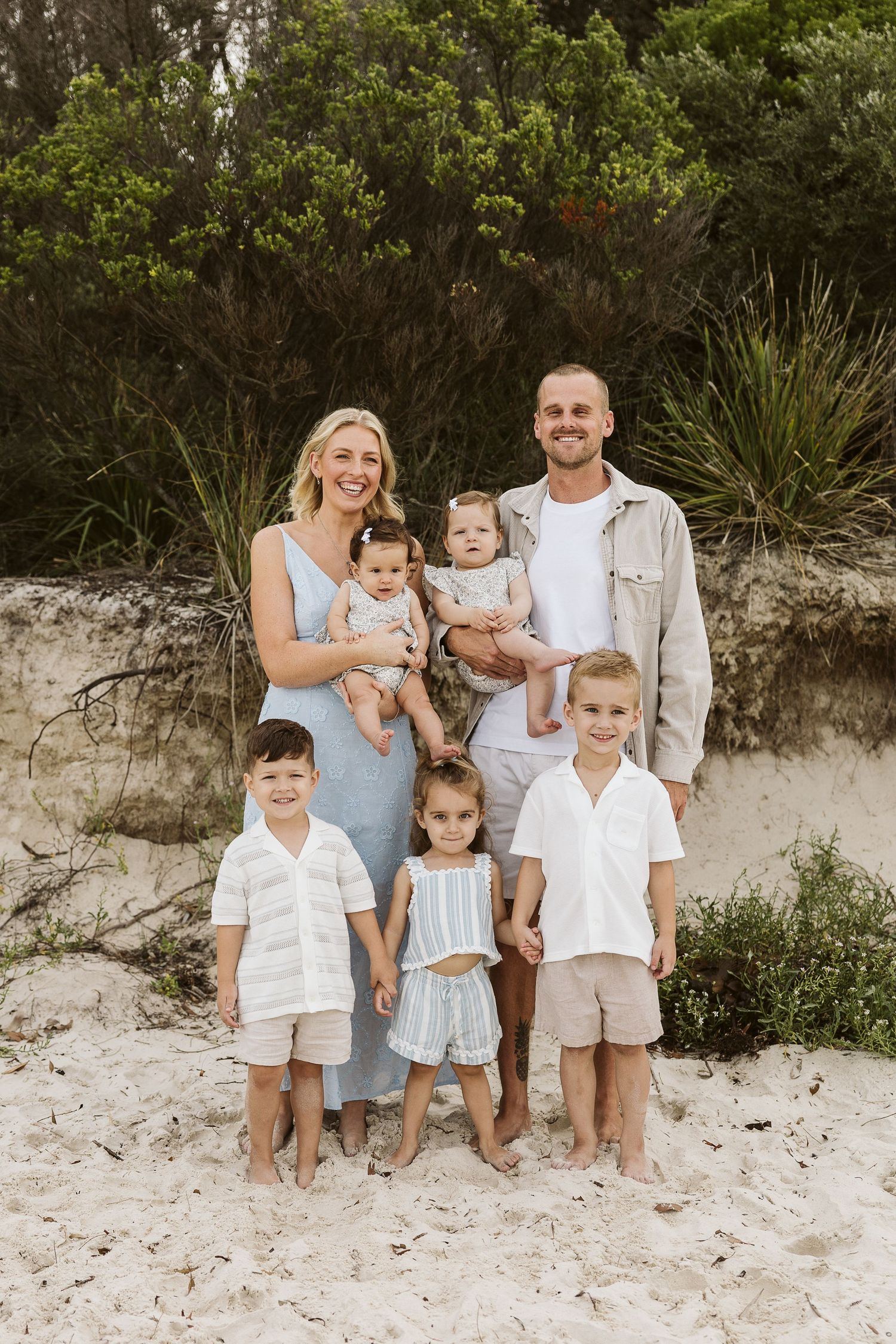 Happy family of seven posing on sandy beach with lush greenery, parents holding two babies with three kids in front.